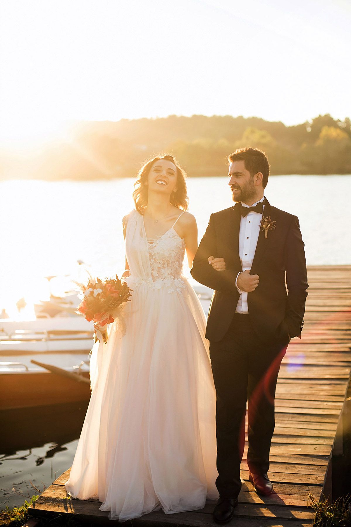A bride and groom walk arm-in-arm down a wooden pier at sunset in Bursa, laughing candidly in a beautiful, sun-drenched bridal portrait.