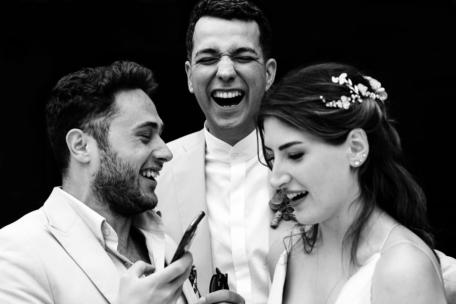 A groom, bride, and friend laugh hysterically together while looking at a phone in this candid black and white Yanık Mektep wedding photo.
