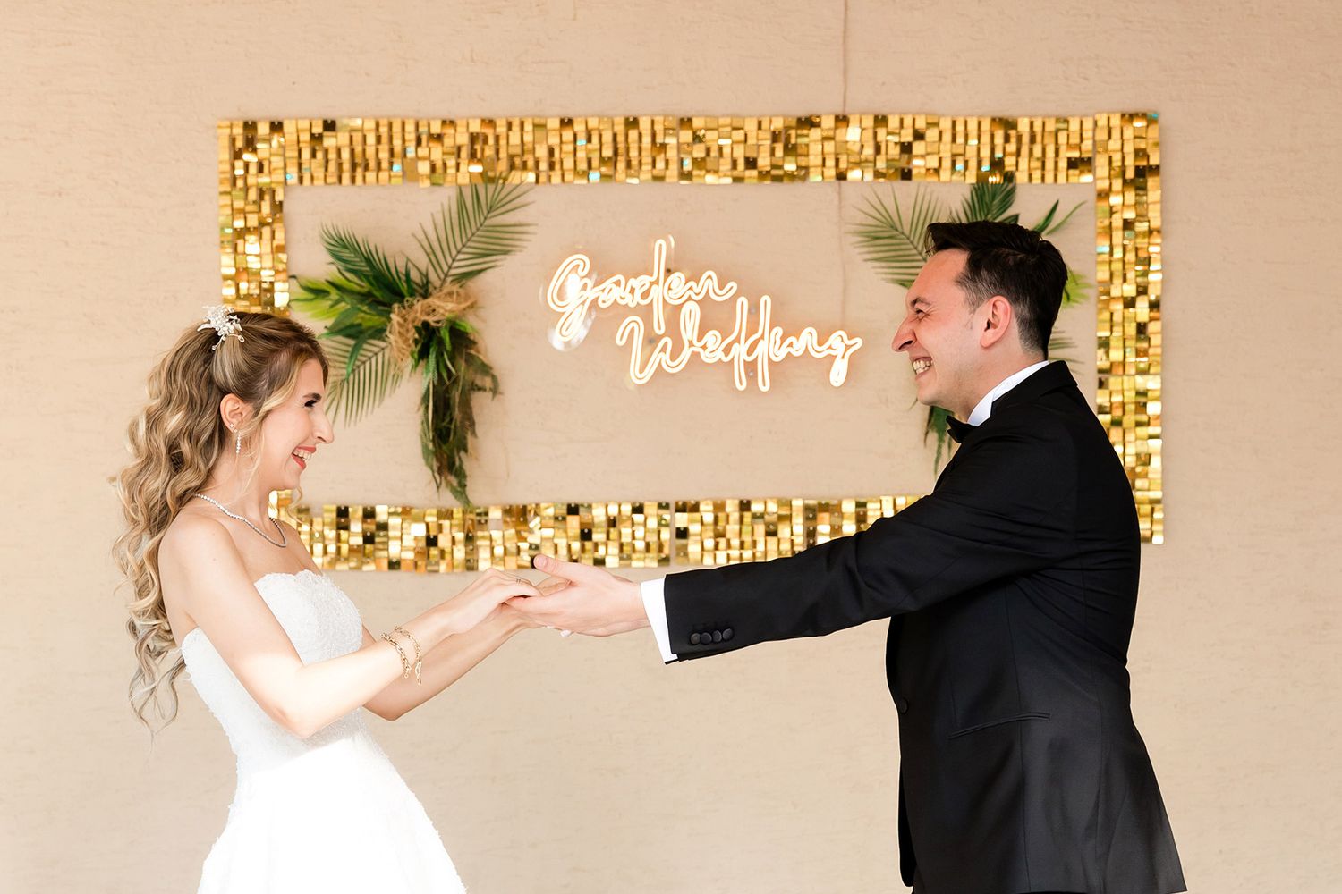 A smiling bride and groom hold hands and laugh in front of a neon 