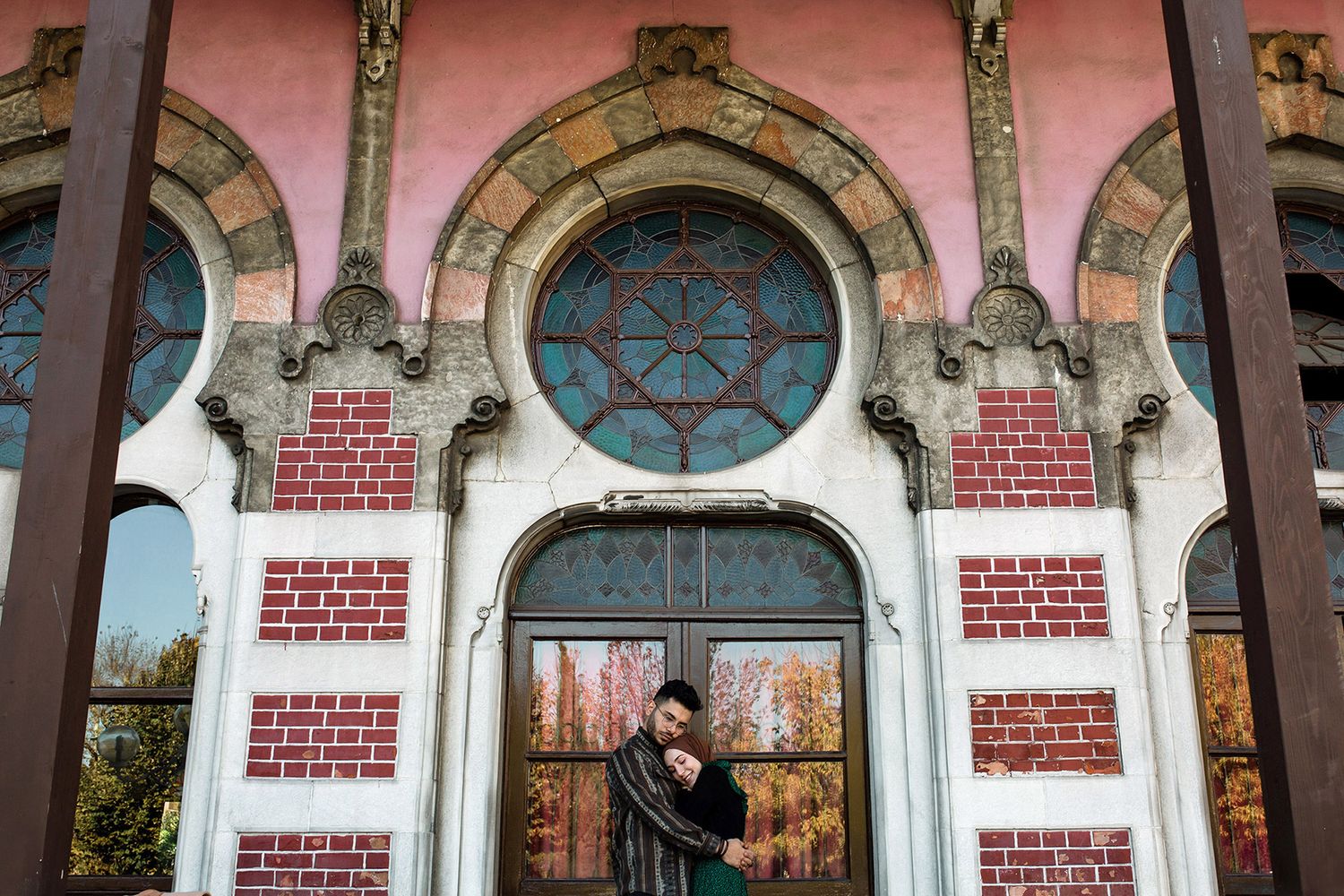 A couple embraces in front of the ornate, historical stained glass windows and pink brick facade of Sirkeci Train Station in Istanbul.