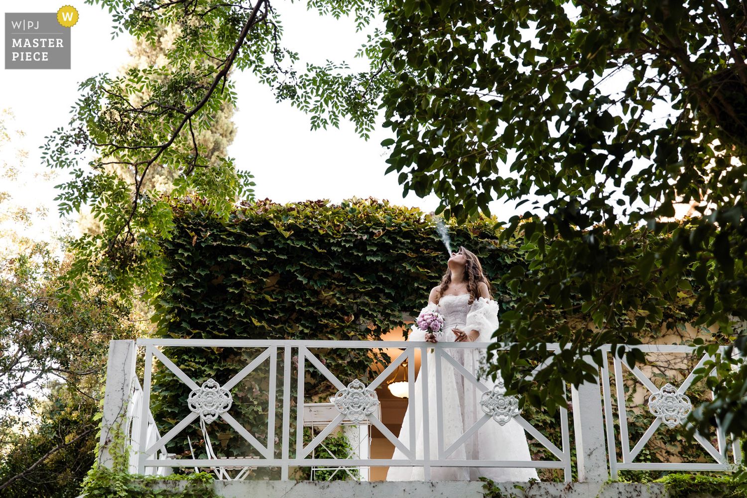 WPJA award-winning documentary portrait of a bride elegantly blowing smoke while standing on a lush, ivy-covered balcony.