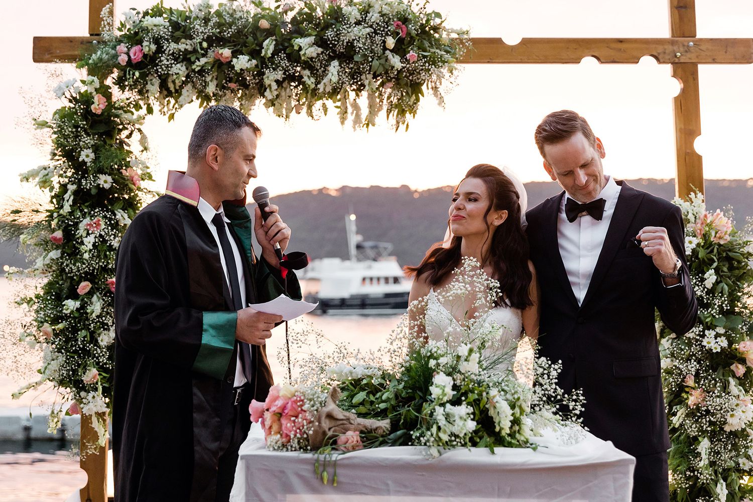Close-up of an officiant in a robe speaking into a microphone during a wedding ceremony at a floral-decorated altar by the Bosphorus strait.