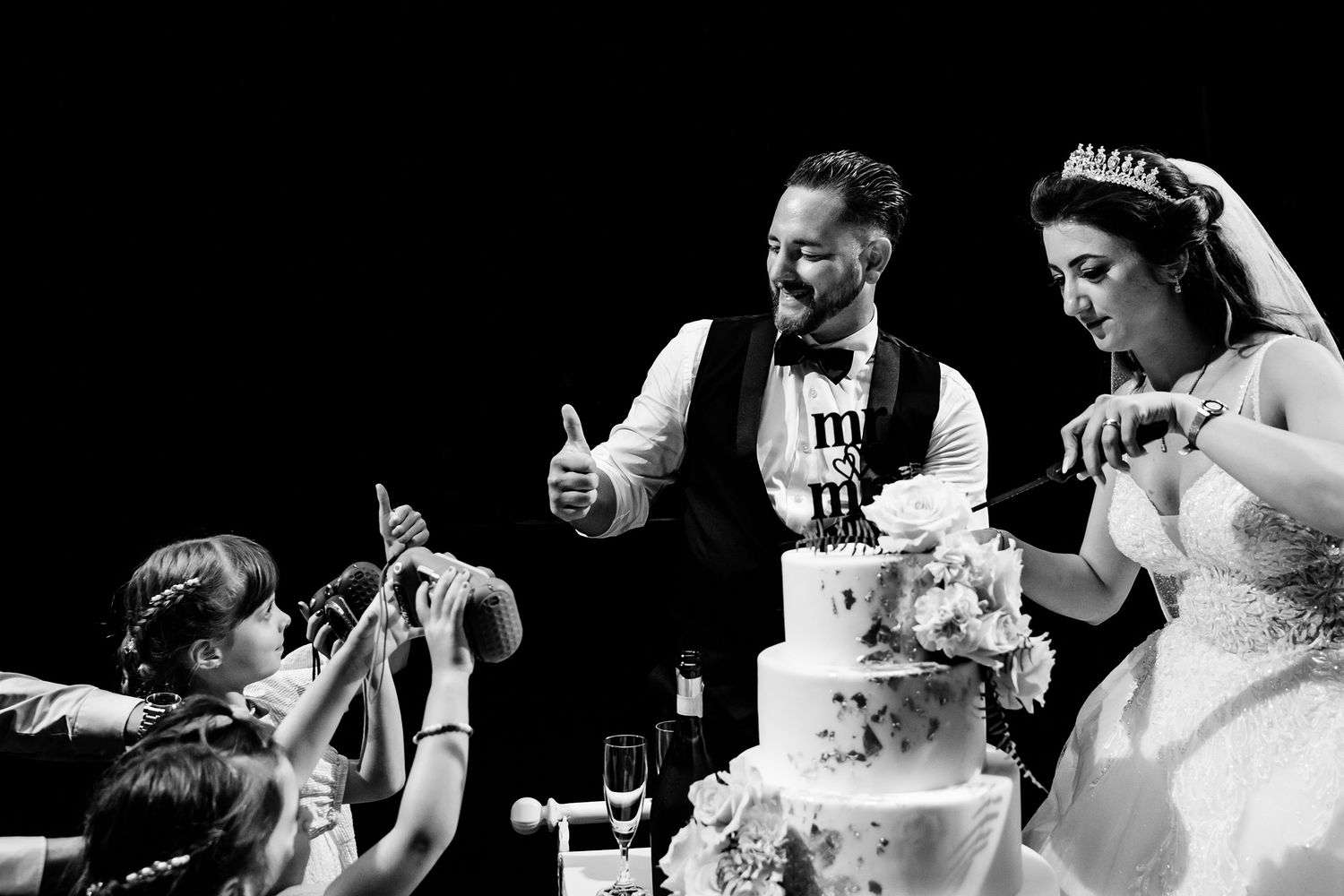 Black and white candid photo of a groom giving a thumbs-up to children taking photos while he and his bride cut their wedding cake.