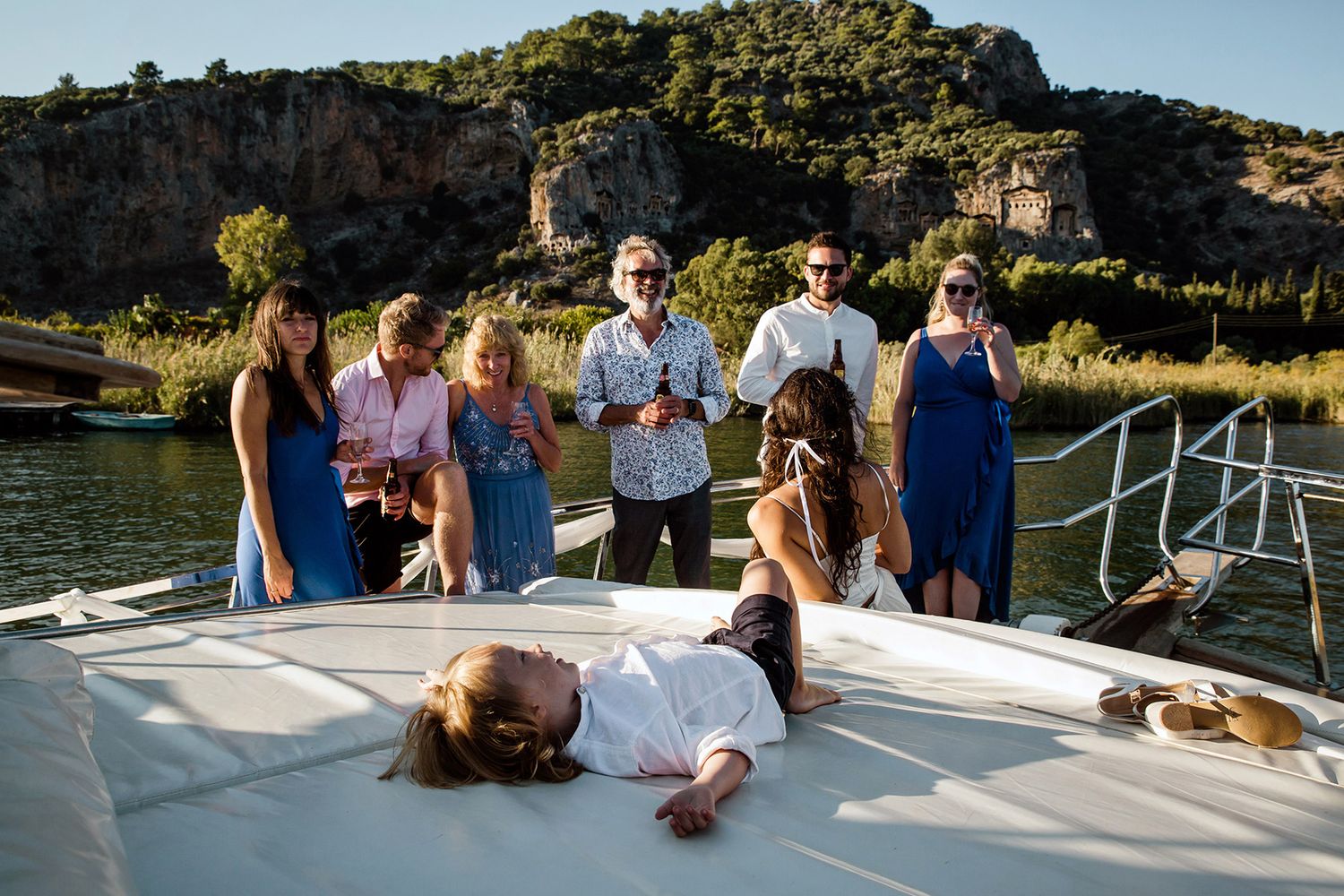 A child sleeps peacefully on a boat deck while adult British guests drink and chat, with the ancient Dalyan rock tombs in the background.