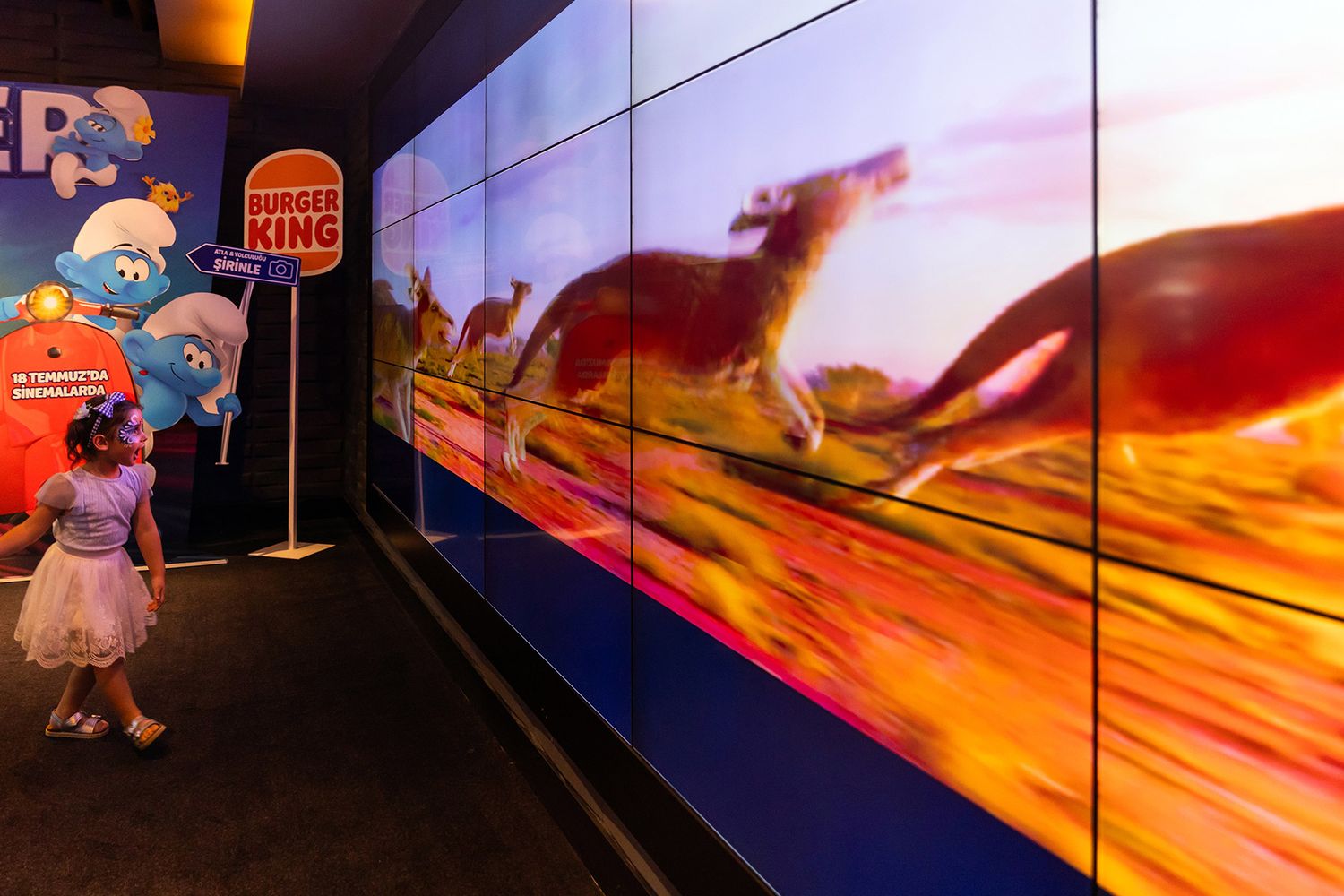 A young girl with face paint walking past a massive digital video wall displaying wildlife next to a Burger King promotional stand.