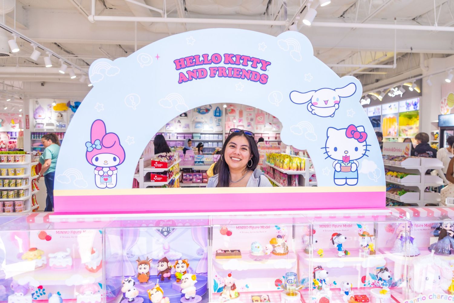 Smiling woman posing at a Hello Kitty and Friends themed display booth inside a Miniso store filled with Sanrio merchandise.