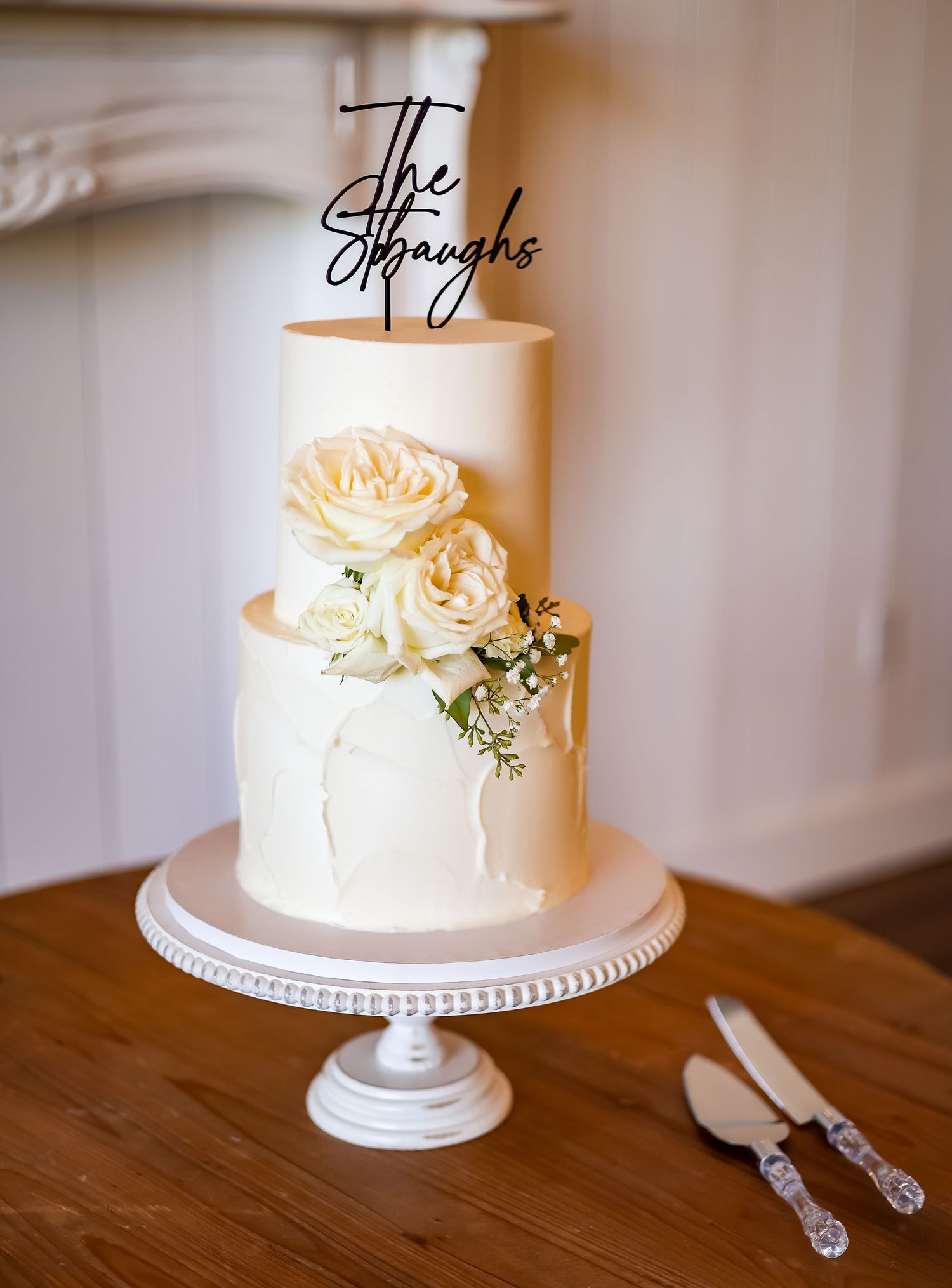 Elegant two-tier white wedding cake with roses and The Straughs topper on a beaded cake stand.