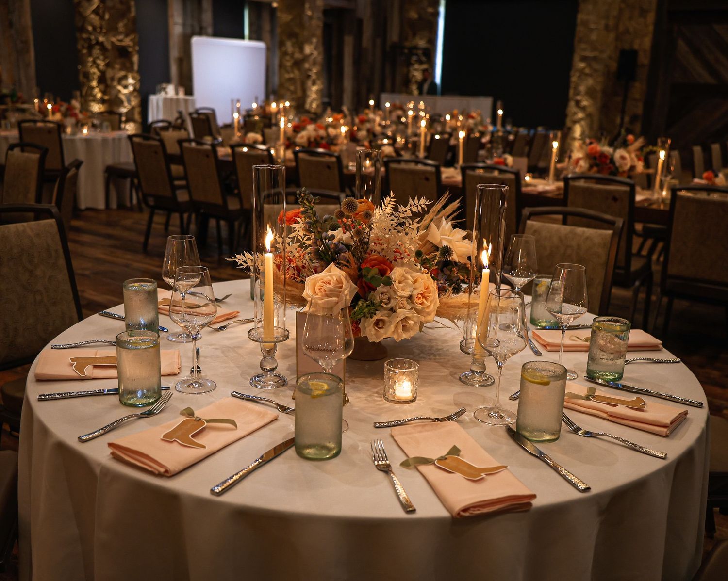 Candlelit round wedding reception table with floral centerpiece, peach napkins, and glassware in a dimly lit venue.