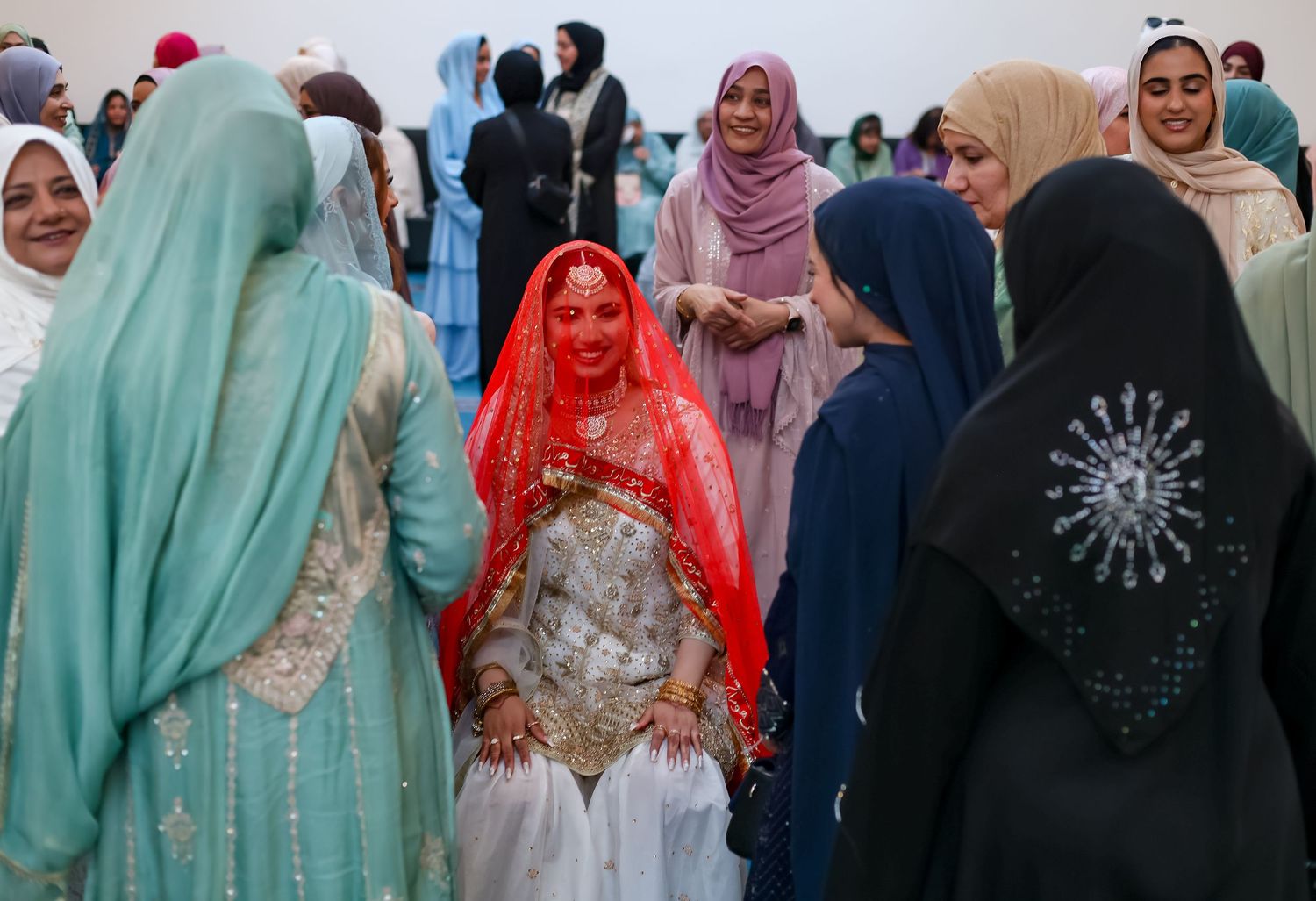 Muslim women in colorful hijabs gather around a bride in a red veil at a traditional Islamic wedding ceremony.