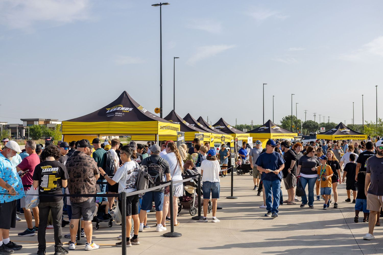 Crowd of people gathered under yellow and black tents at an outdoor event in a sunny parking lot.