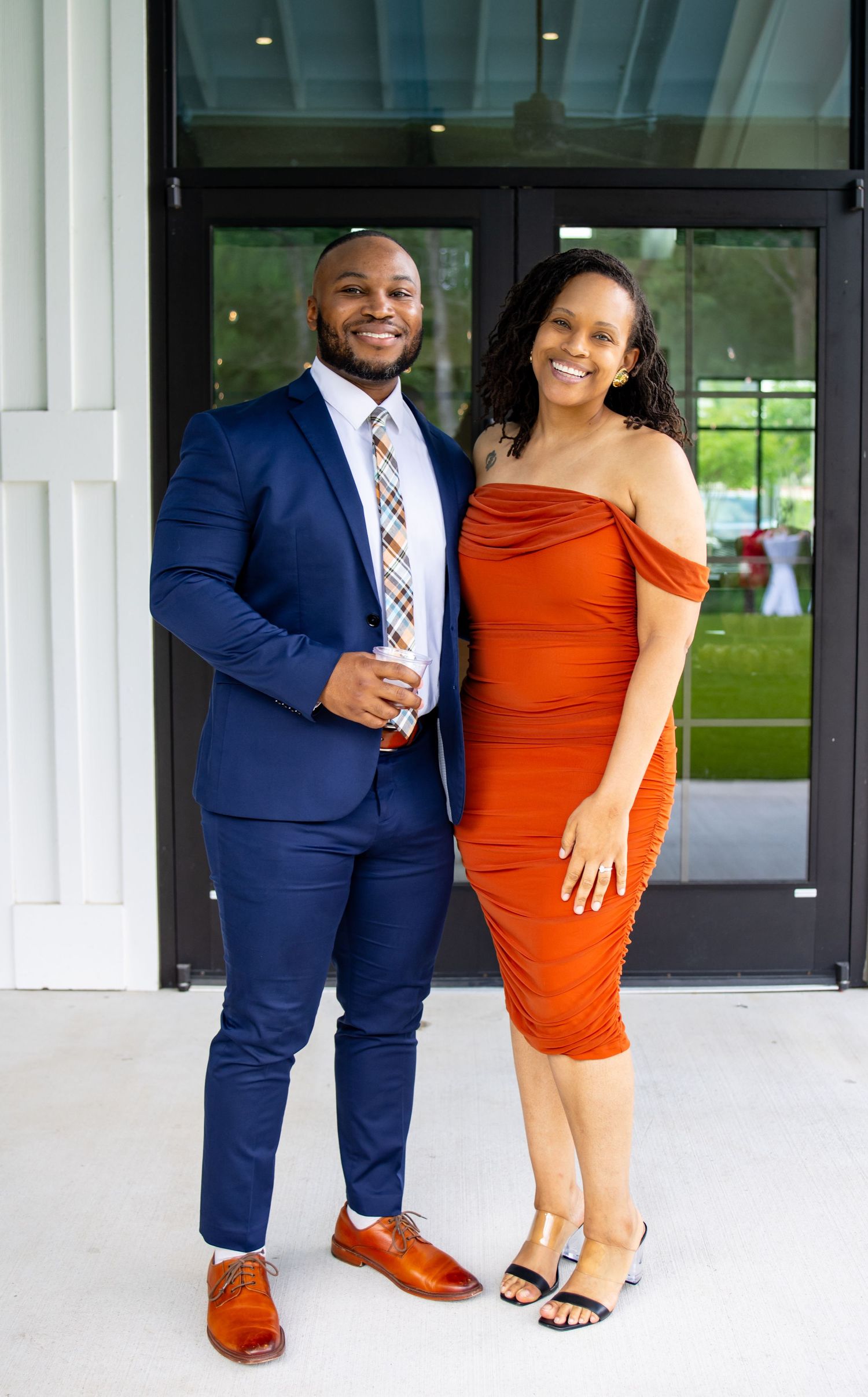 Couple posing outdoors, man in navy suit with colorful tie, woman in rust off-shoulder dress, smiling at formal event.