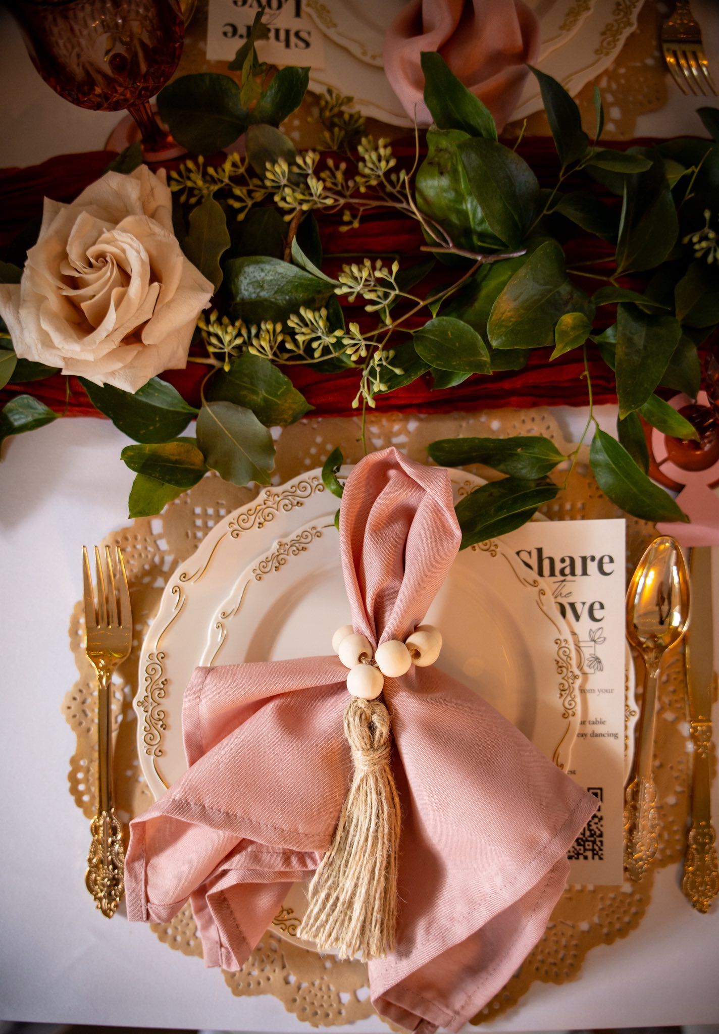 Elegant wedding place setting with dusty rose napkin, gold cutlery, tassel ring, and floral greenery centerpiece.