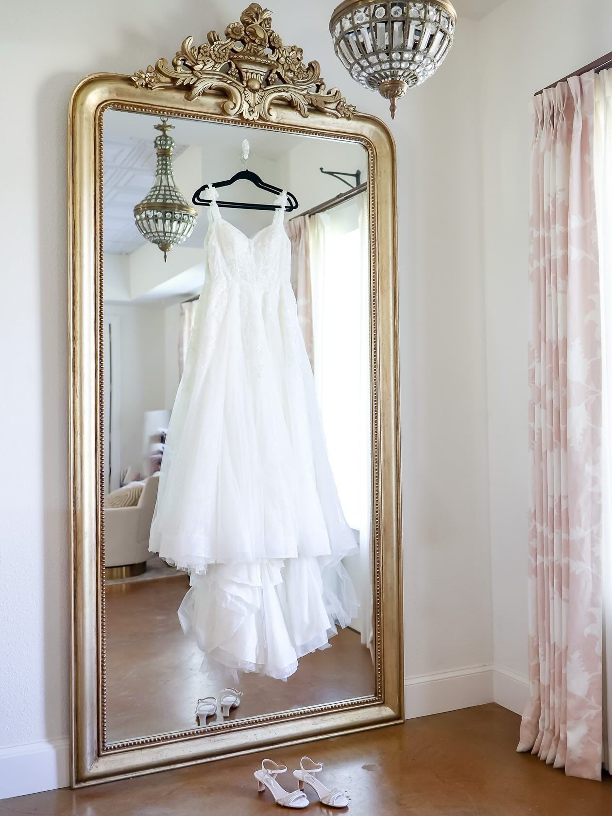 Elegant white A-line wedding dress hanging on an ornate gold floor mirror with crystal chandelier reflection.