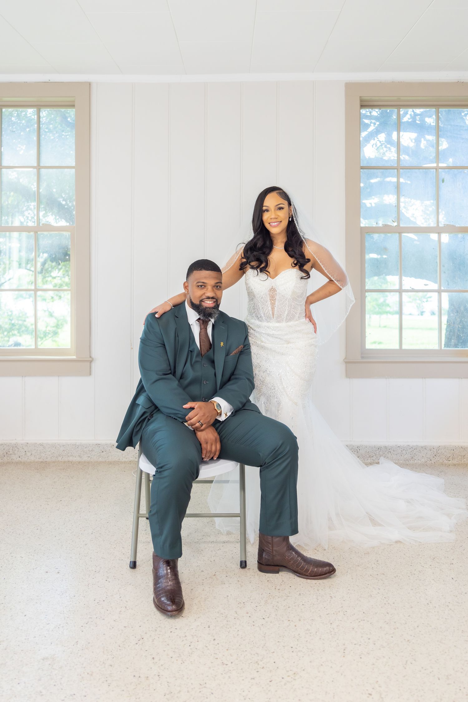 Bride in lace mermaid gown and groom in green suit posing elegantly in bright white room.