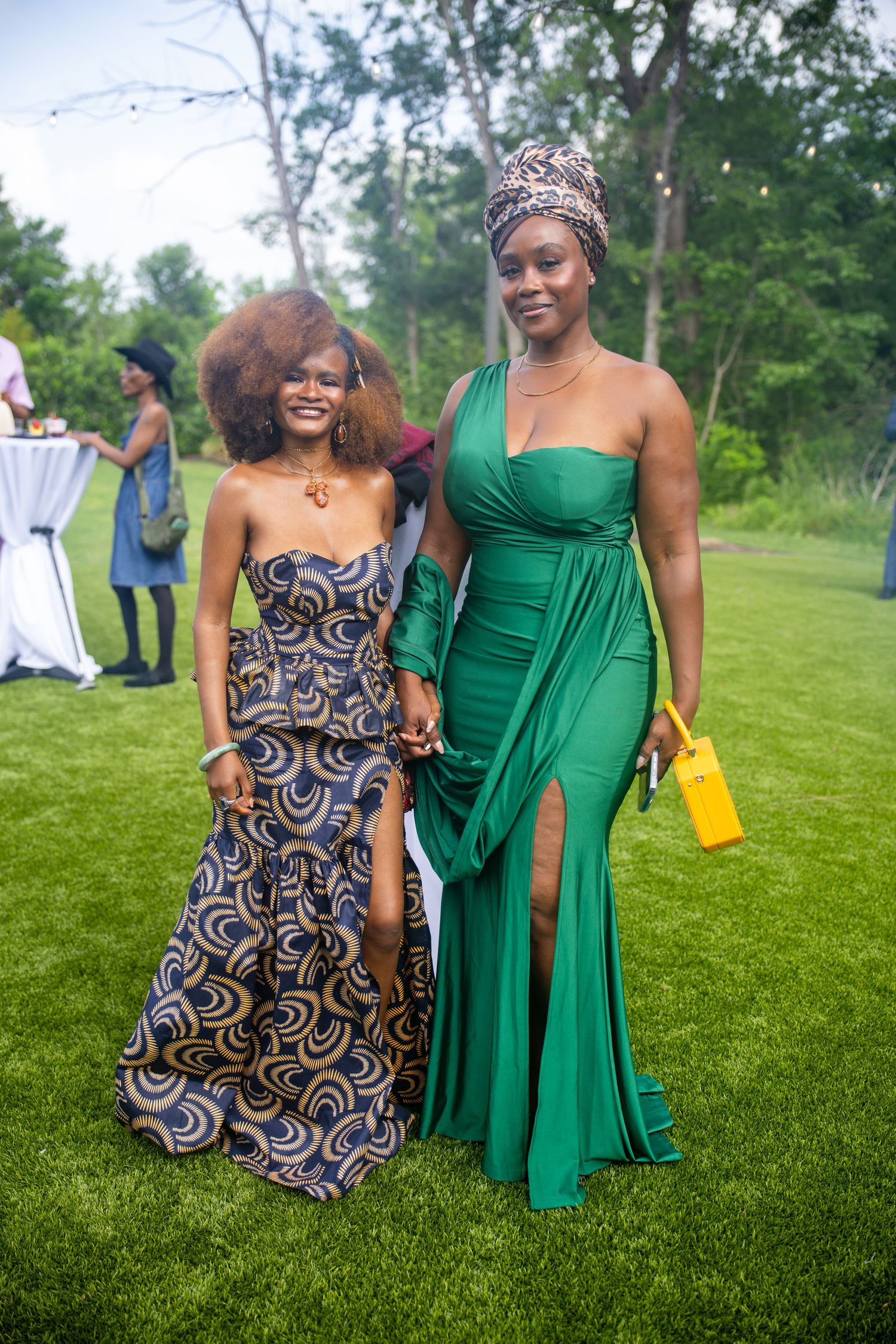 Two elegantly dressed women posing outdoors at a garden event, one in a patterned dress and one in a green gown.