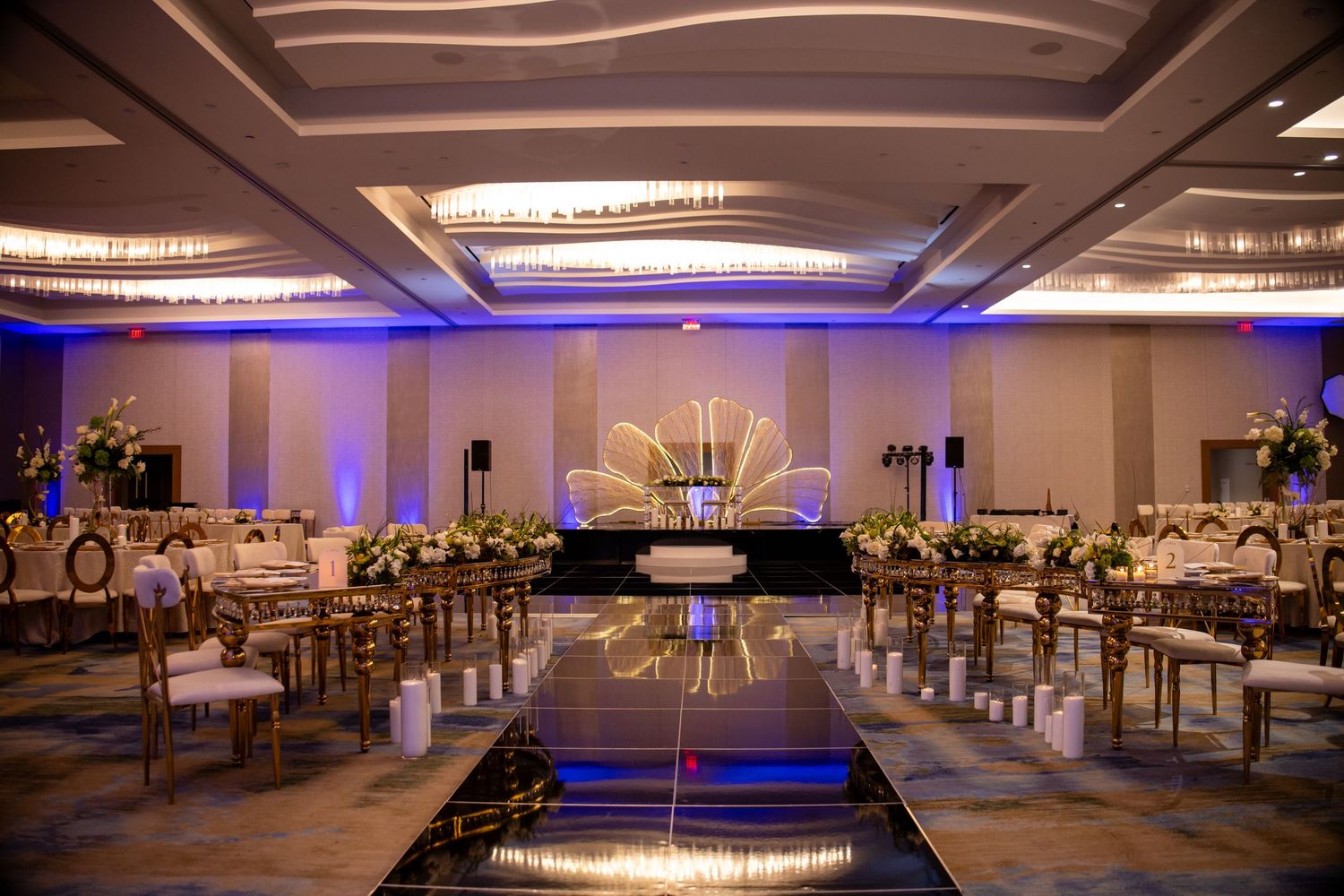 Elegant ballroom wedding reception with gold chairs, floral centerpieces, mirrored aisle, and blue uplighting.