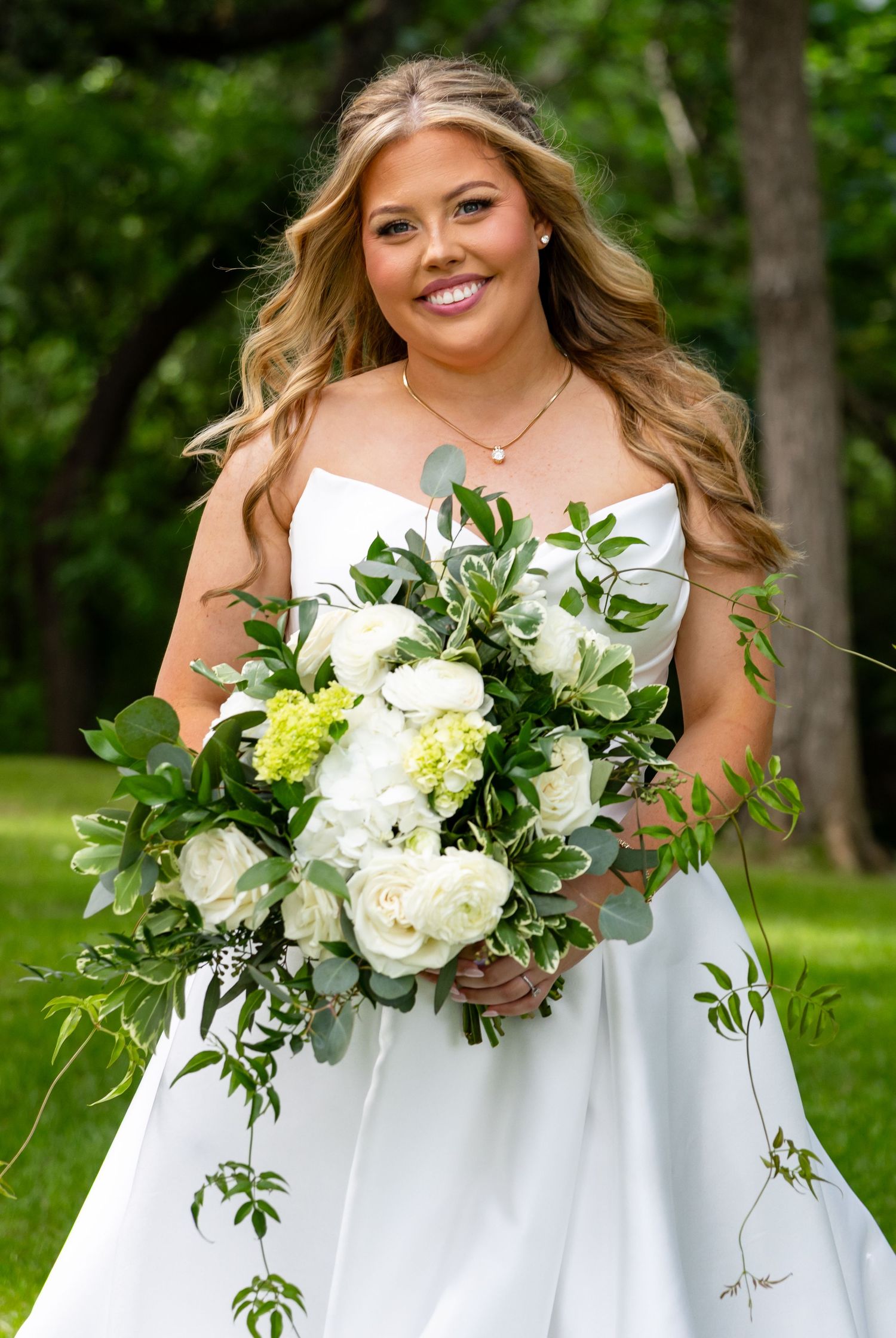 Glowing bride holds a stunning white rose and greenery bouquet while smiling outdoors in a strapless gown.