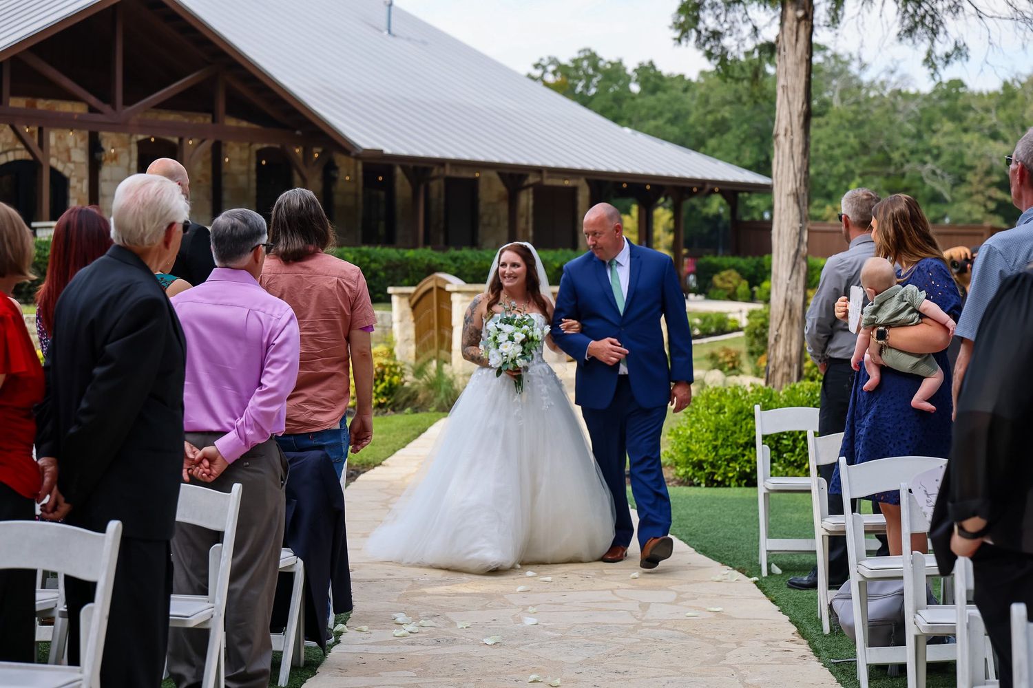 Bride in ball gown walks down outdoor aisle with escort past seated wedding guests.