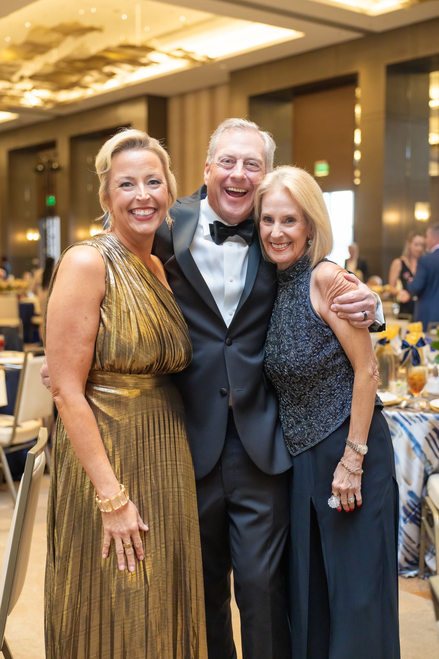 Two women and a man in formal attire smile together at an elegant gala event.
