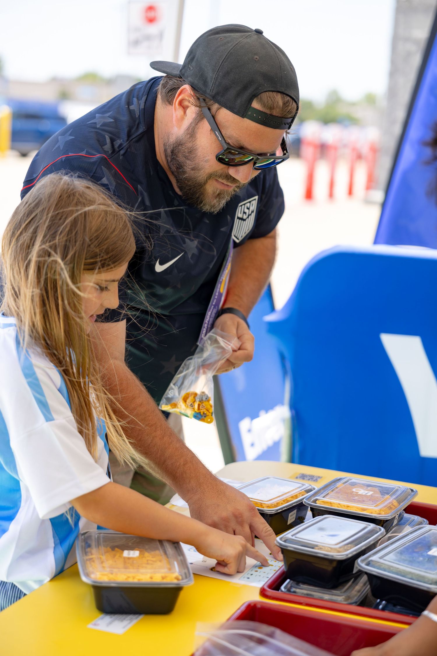 Father and daughter sorting yellow LEGO bricks together at an outdoor building activity table.