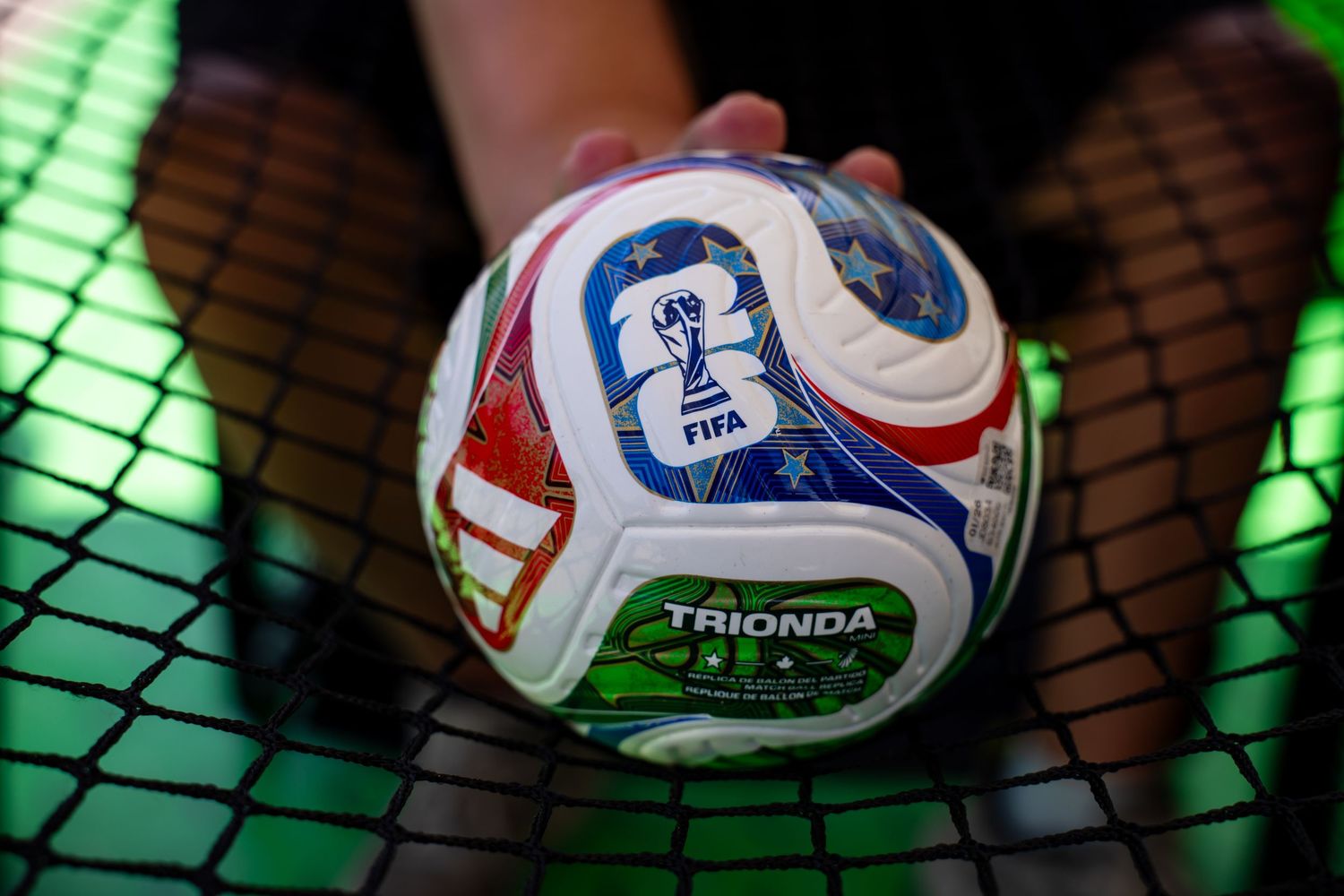 A hand holds a FIFA Trionda official soccer ball against a green net background.