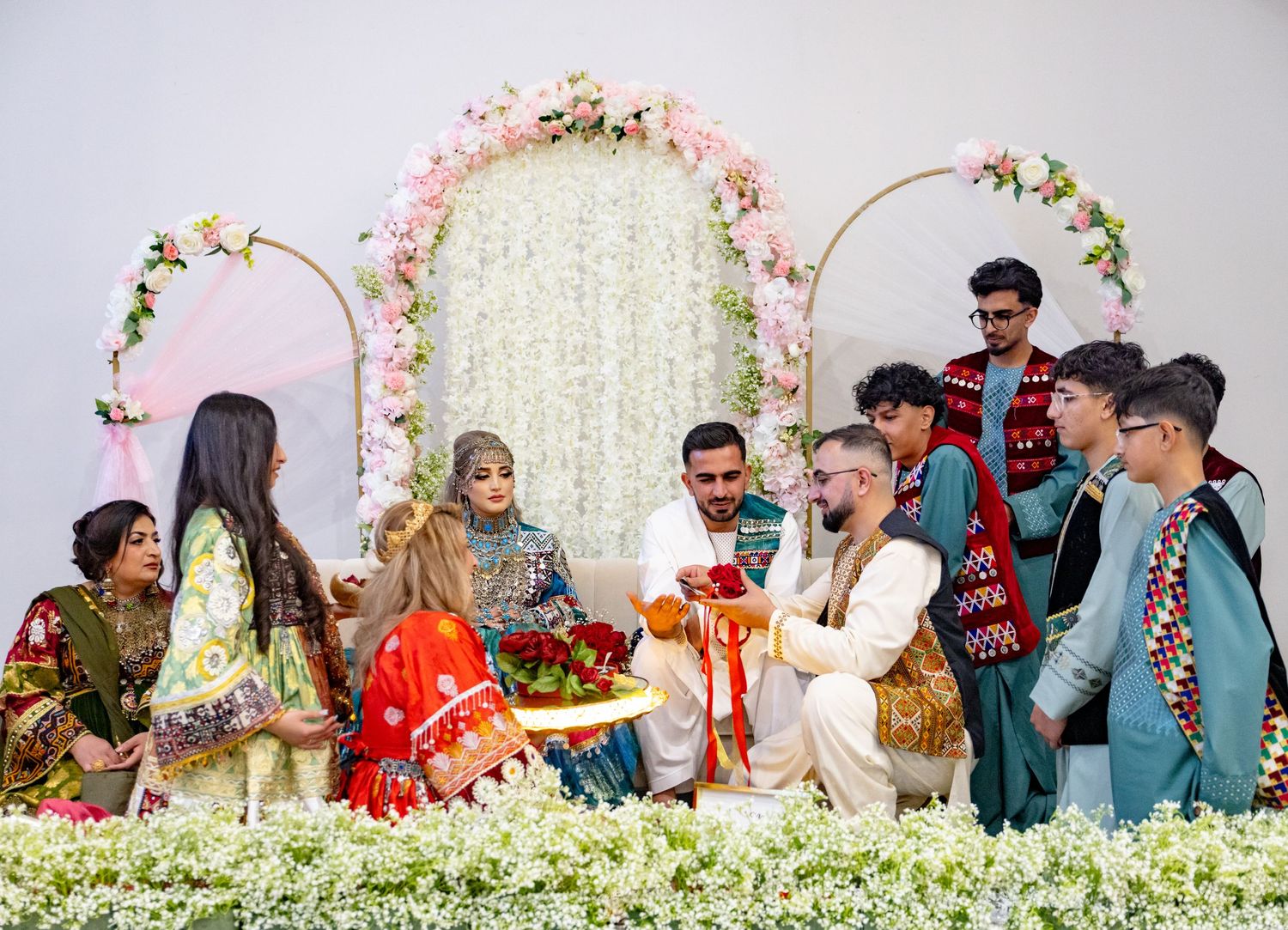 A vibrant South Asian wedding ceremony with guests in colorful traditional attire gathered around the couple near a floral arch backdrop.