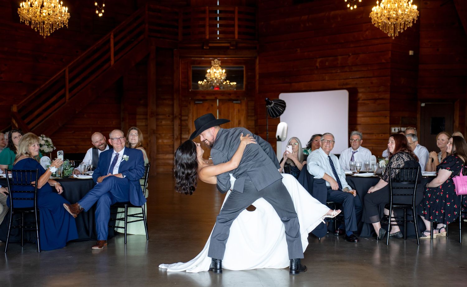 Groom dips bride dramatically on the dance floor as wedding guests watch and applaud in rustic barn.
