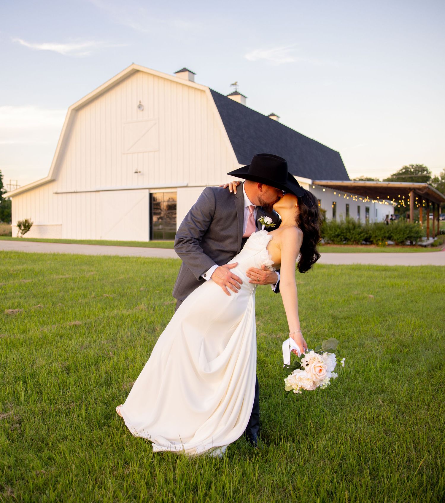 Bride and groom share a romantic dip kiss in front of a white barn at a rustic outdoor wedding venue.