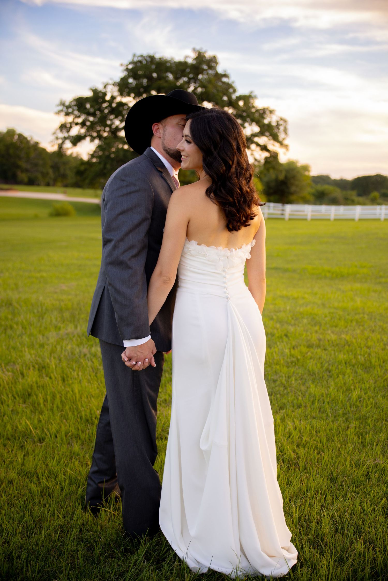 Bride and groom hold hands in a golden sunset field, groom in cowboy hat leaning in for a kiss.