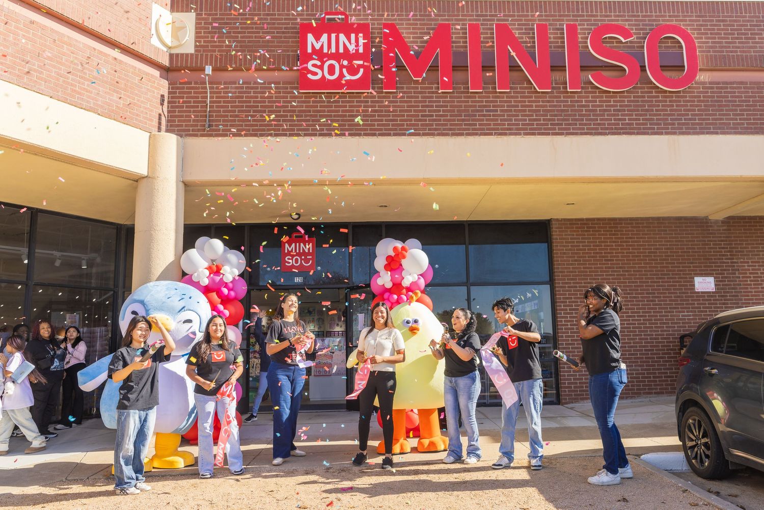 Miniso store grand opening celebration with confetti, mascots, and staff gathered outside the brick storefront entrance.