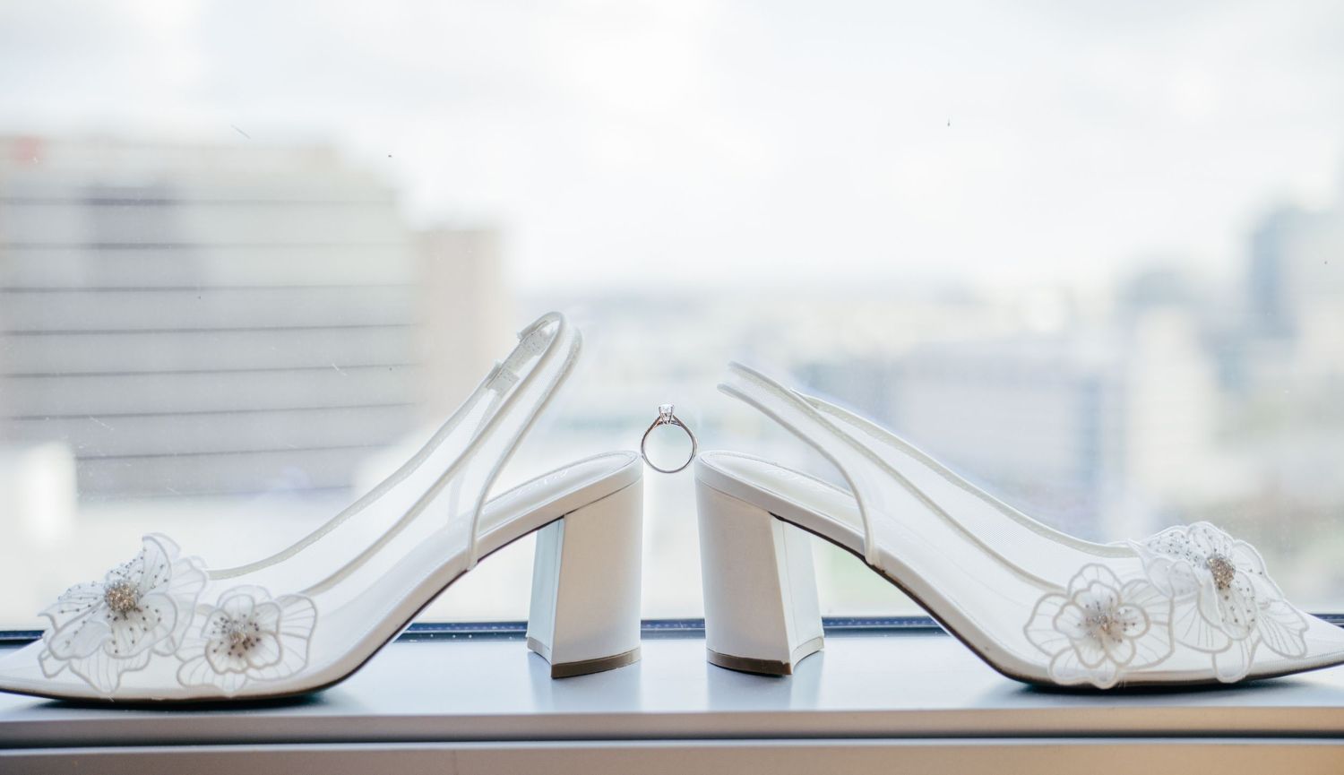 White embellished bridal heels with diamond ring balanced between them on windowsill.