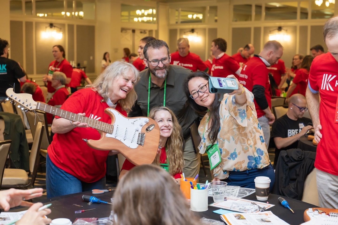 Group of people in red shirts laughing and posing with a guitar at an indoor team building event.