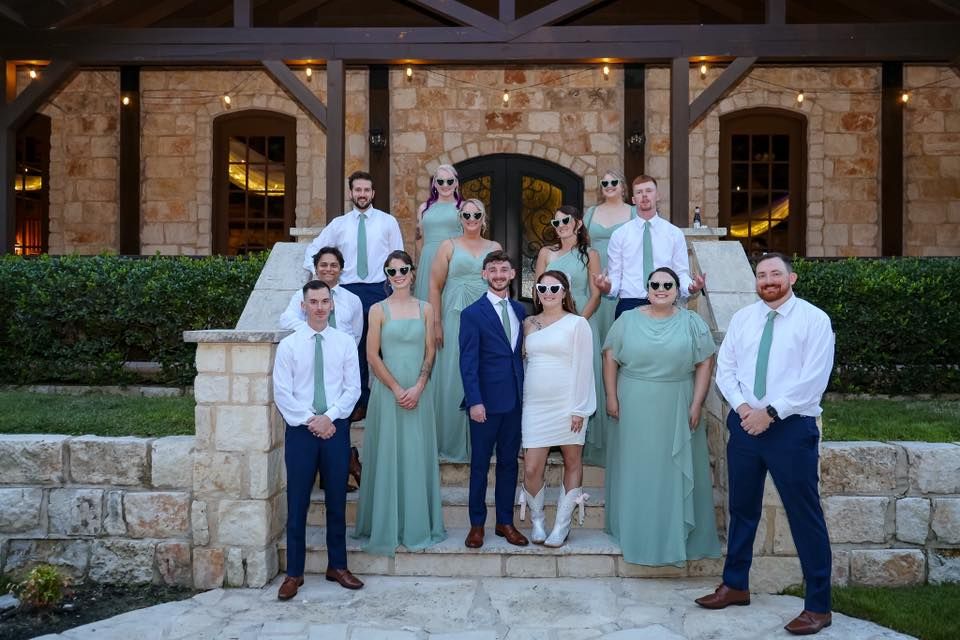 Wedding party poses in sage green and navy attire on stone steps outside an elegant venue.
