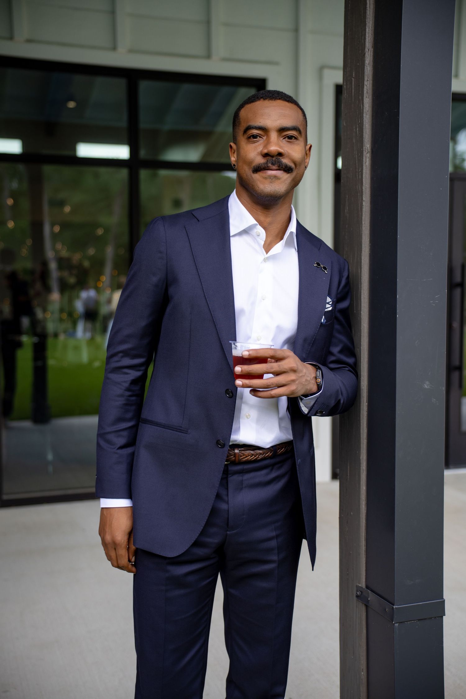 Well-dressed man in navy suit holding a cocktail glass, standing outdoors near a modern building.