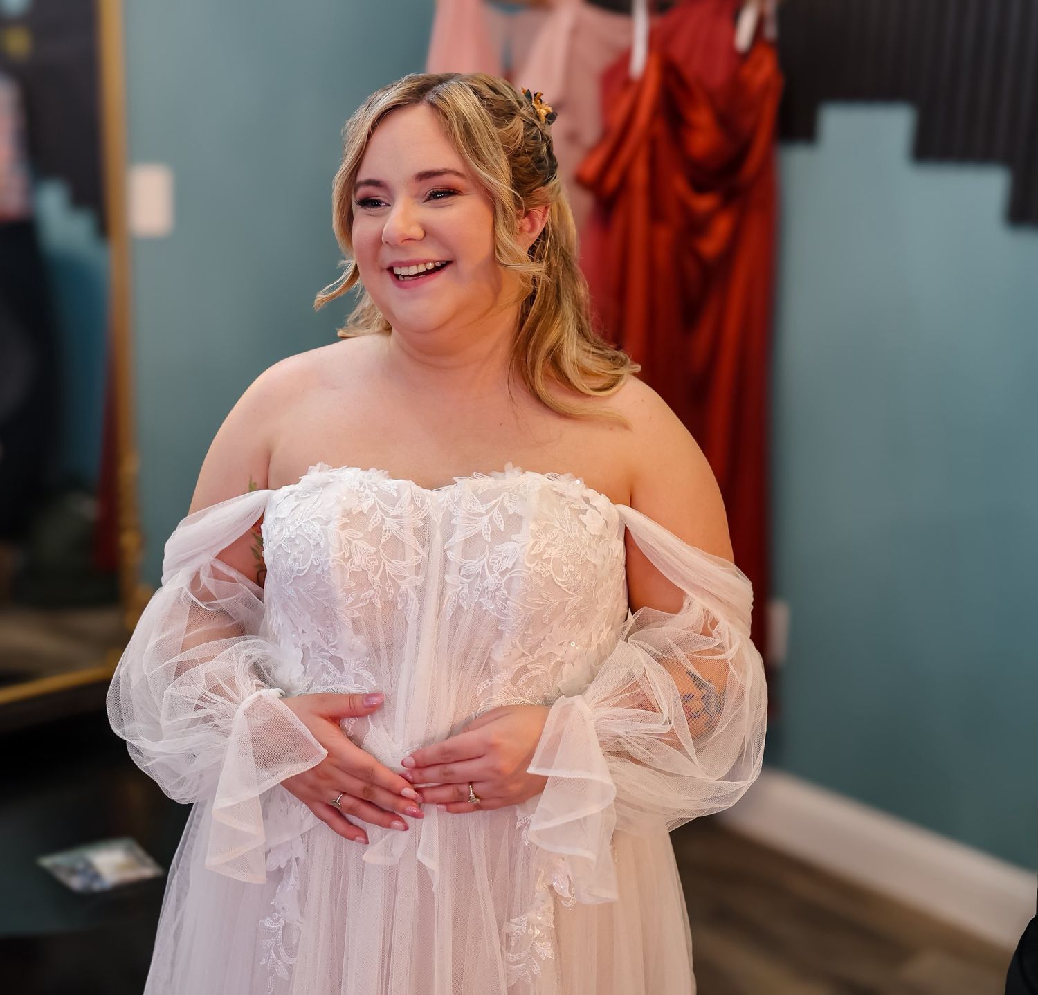 Happy bride trying on off-shoulder lace wedding gown with sheer puff sleeves in bridal boutique.