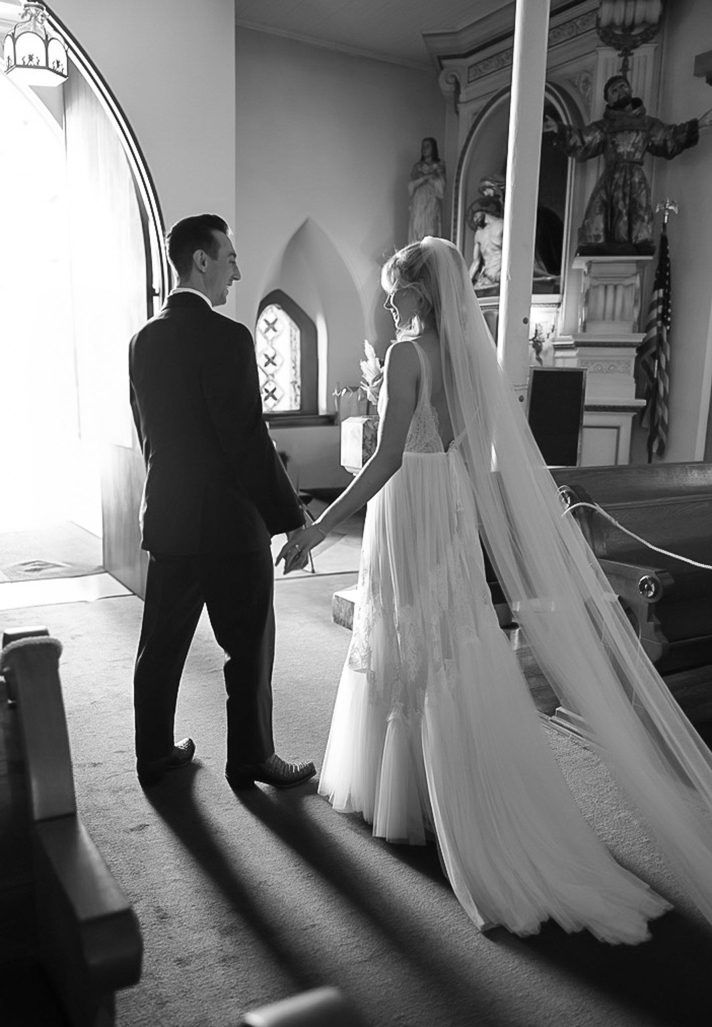 Black and white photo of bride and groom holding hands in a sunlit church with stained glass windows.