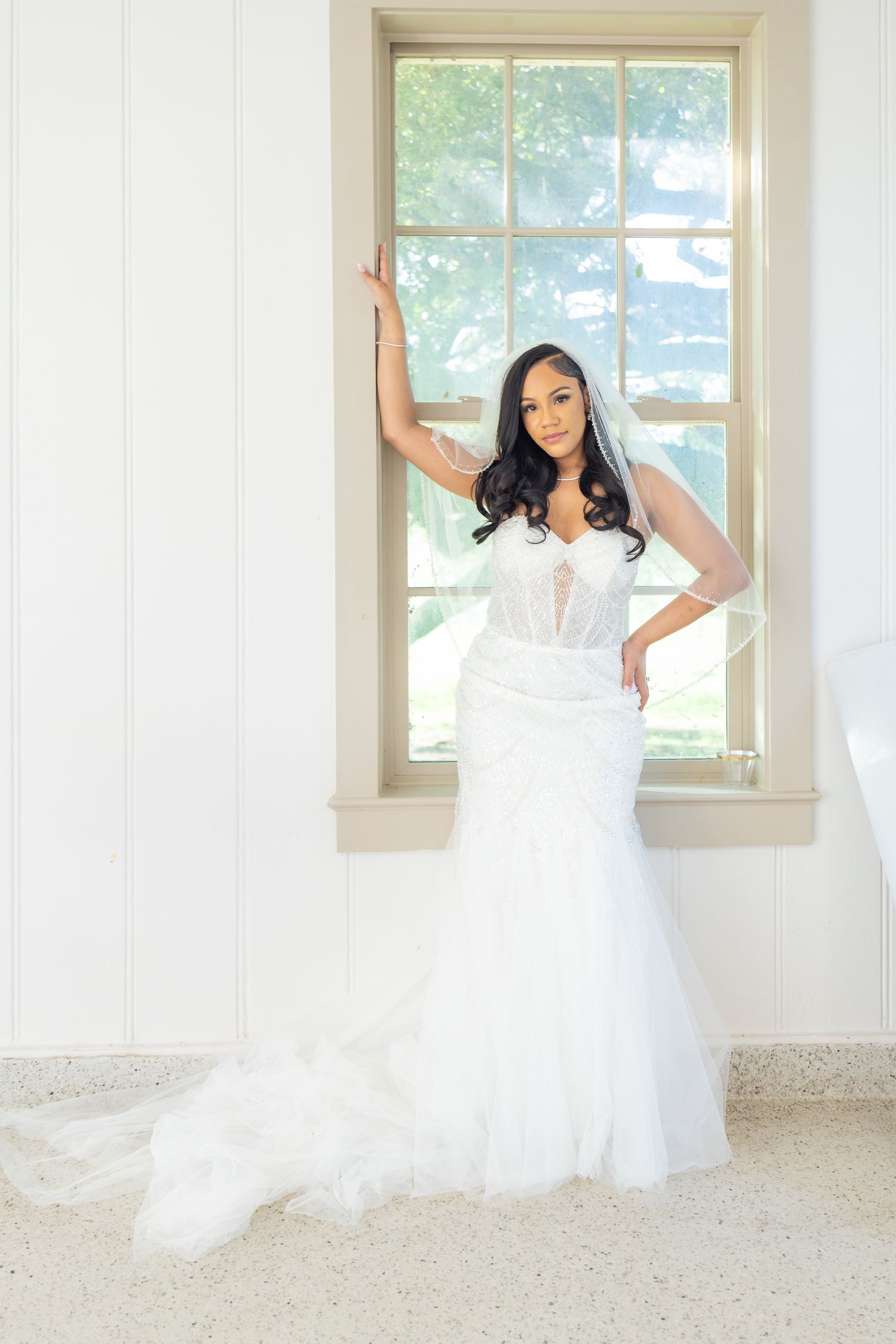 Bride in white mermaid wedding gown with veil posing gracefully by window in bright white room.