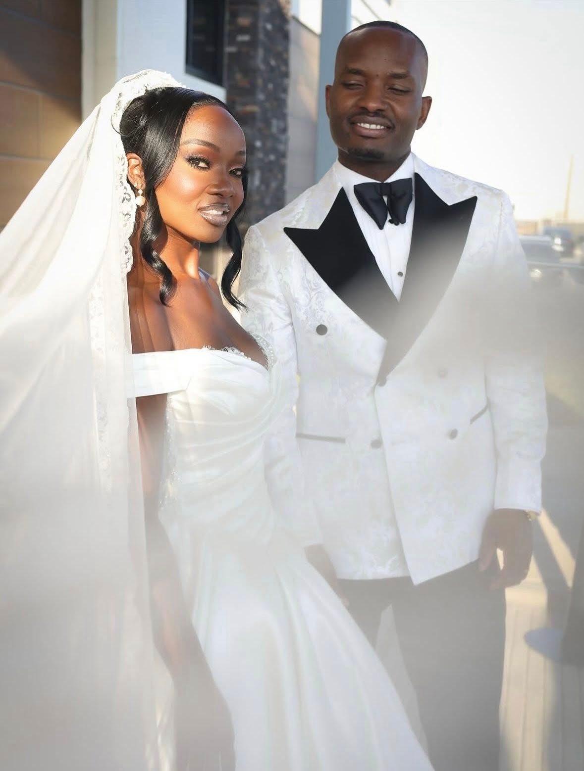 African American bride in white strapless gown with veil poses with groom in white tuxedo at wedding.