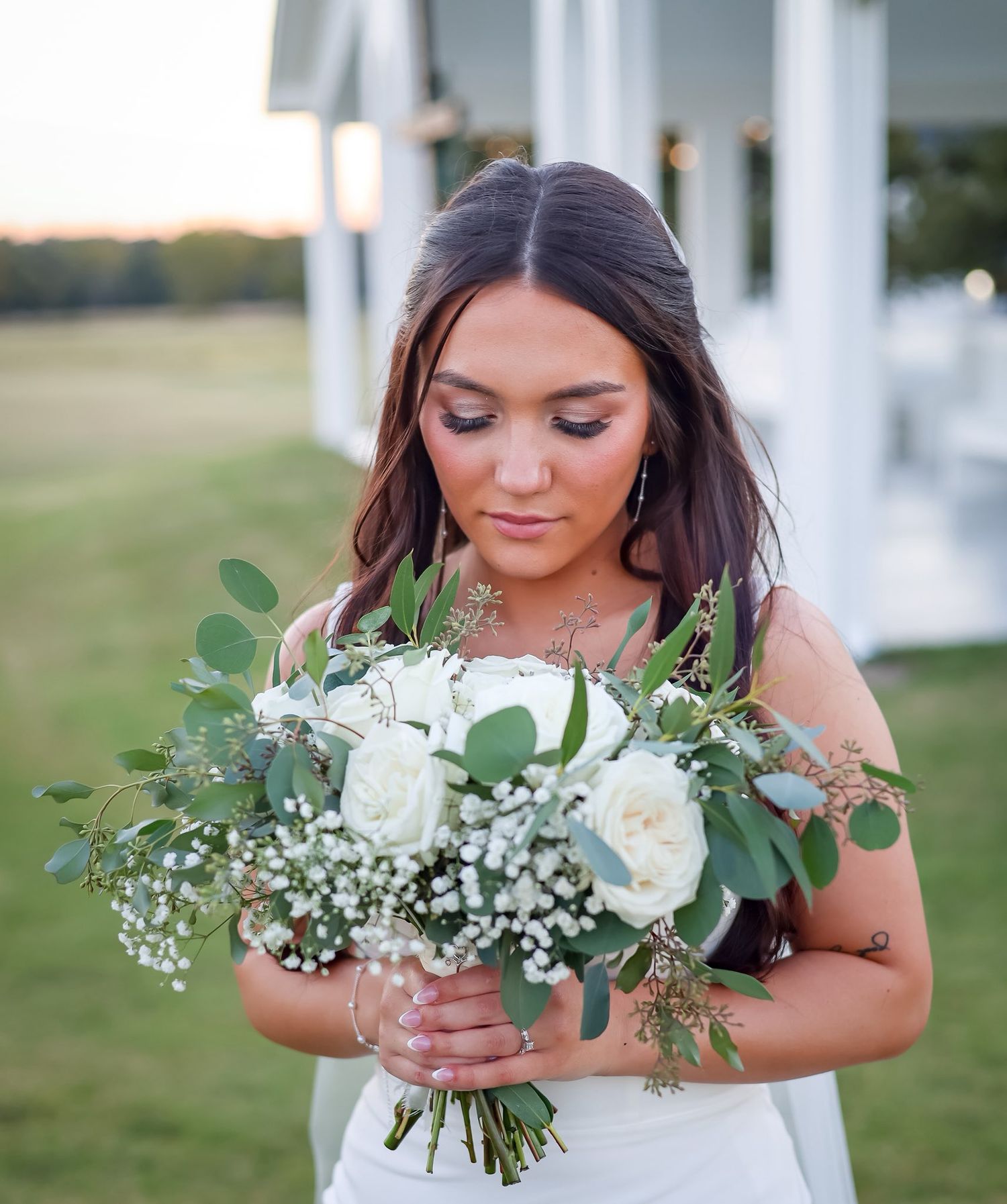 Beautiful bride holding white rose and eucalyptus bouquet in a sleeveless wedding gown at sunset.