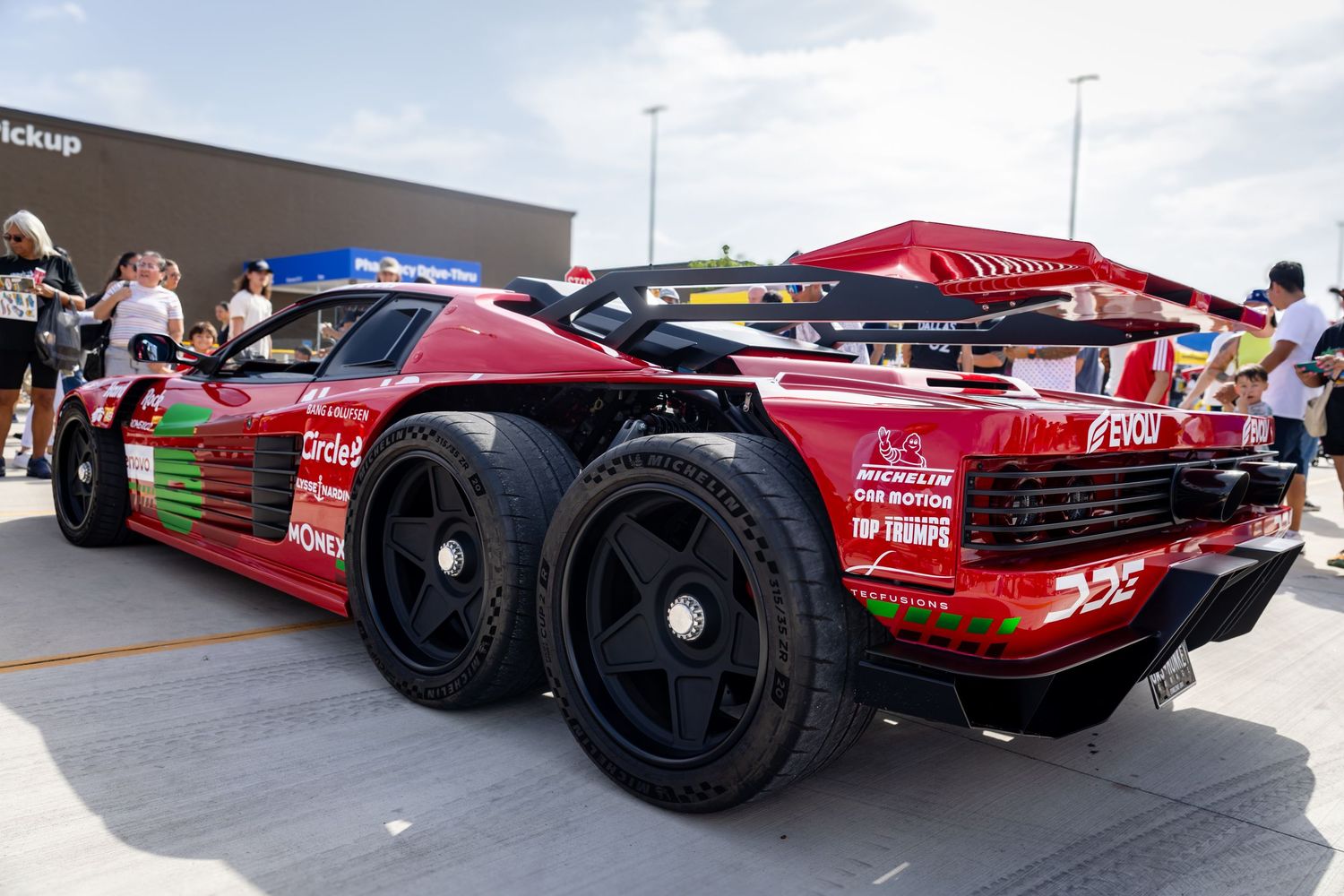 Red Ferrari 6-wheel custom race car displayed at an outdoor motorsport event with spectators in the background.
