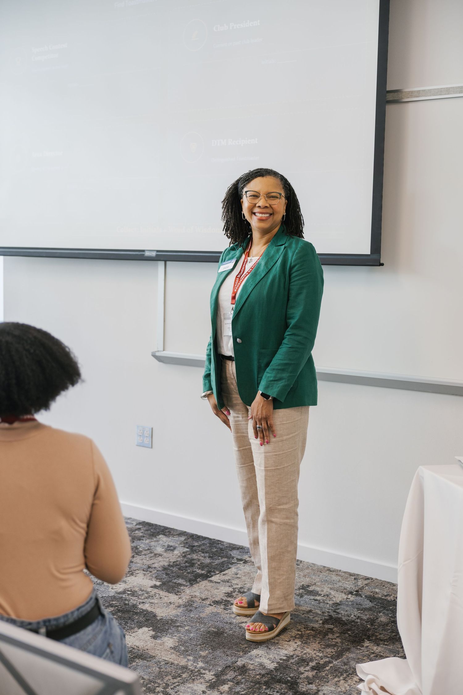 A smiling woman in a green blazer and glasses stands before a whiteboard in a classroom setting.