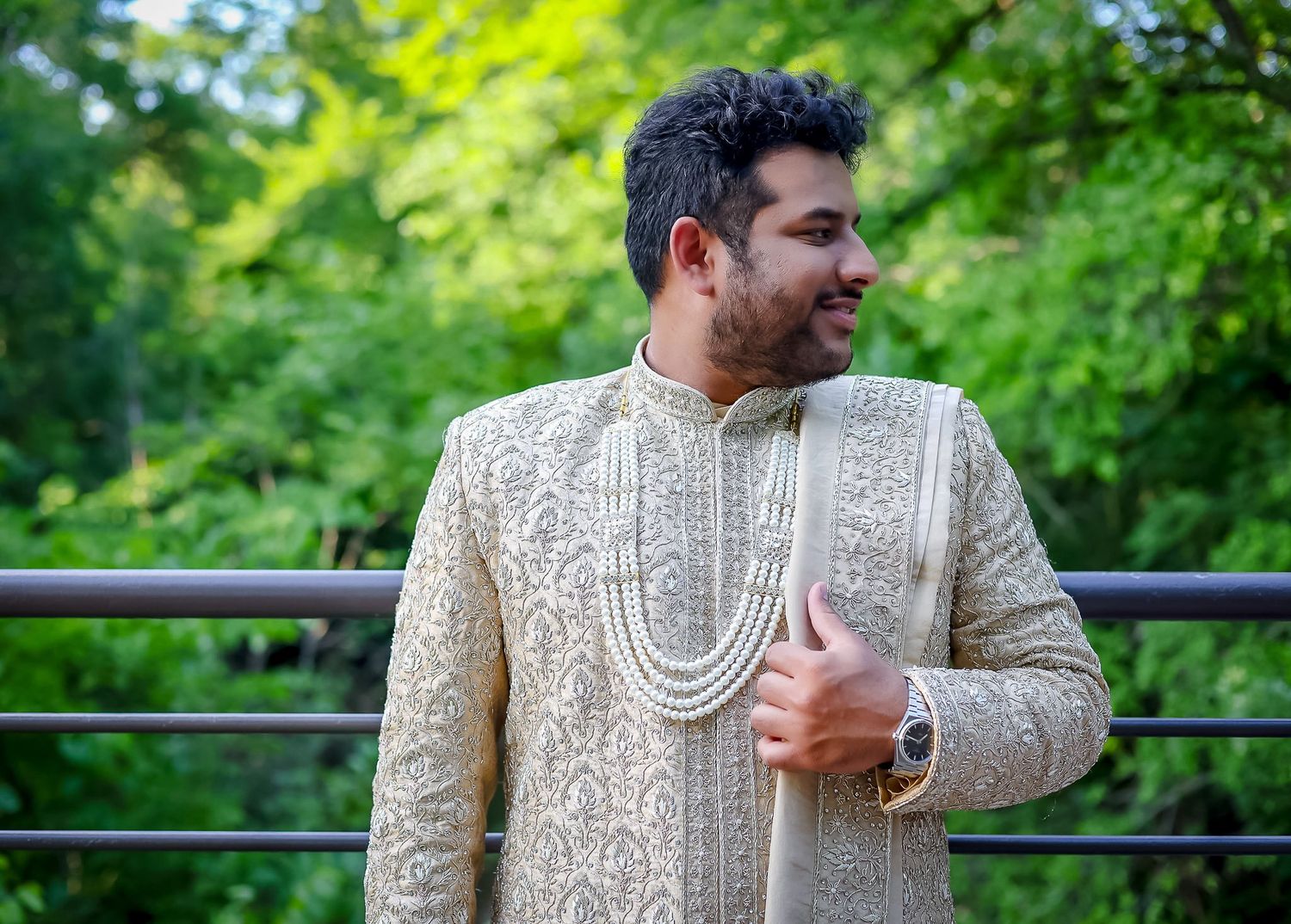 Man in ornate gold sherwani with pearl necklace posing outdoors on bridge surrounded by lush green trees.