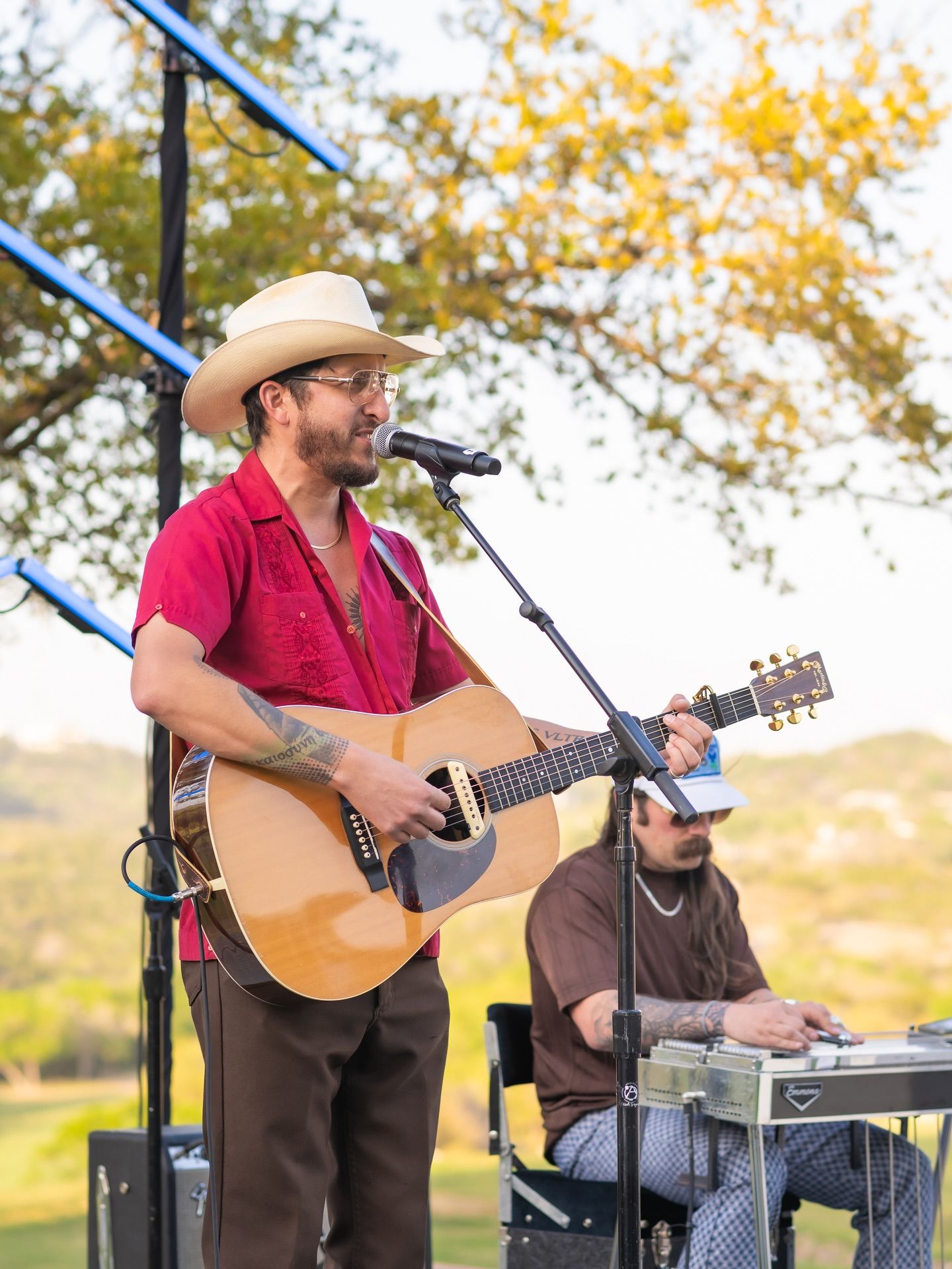 Country musician in red shirt and straw hat playing acoustic guitar on an outdoor stage at sunset.