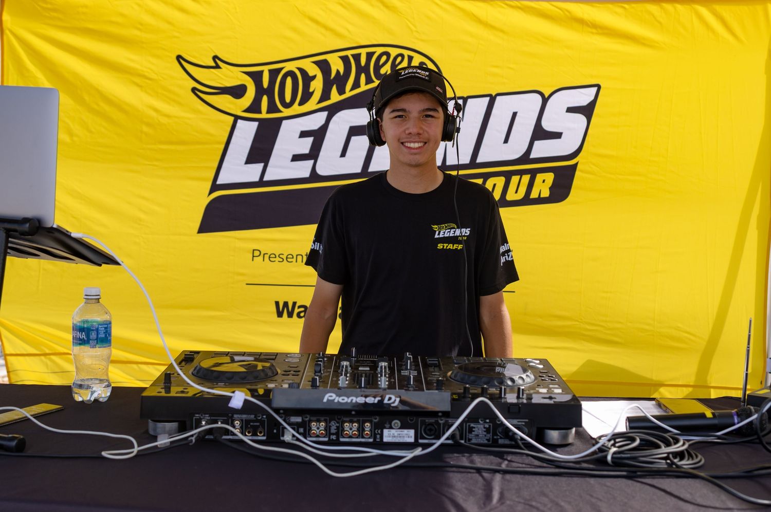 A young DJ smiles behind turntables at the Hot Wheels Legends Tour event with a yellow branded banner backdrop.