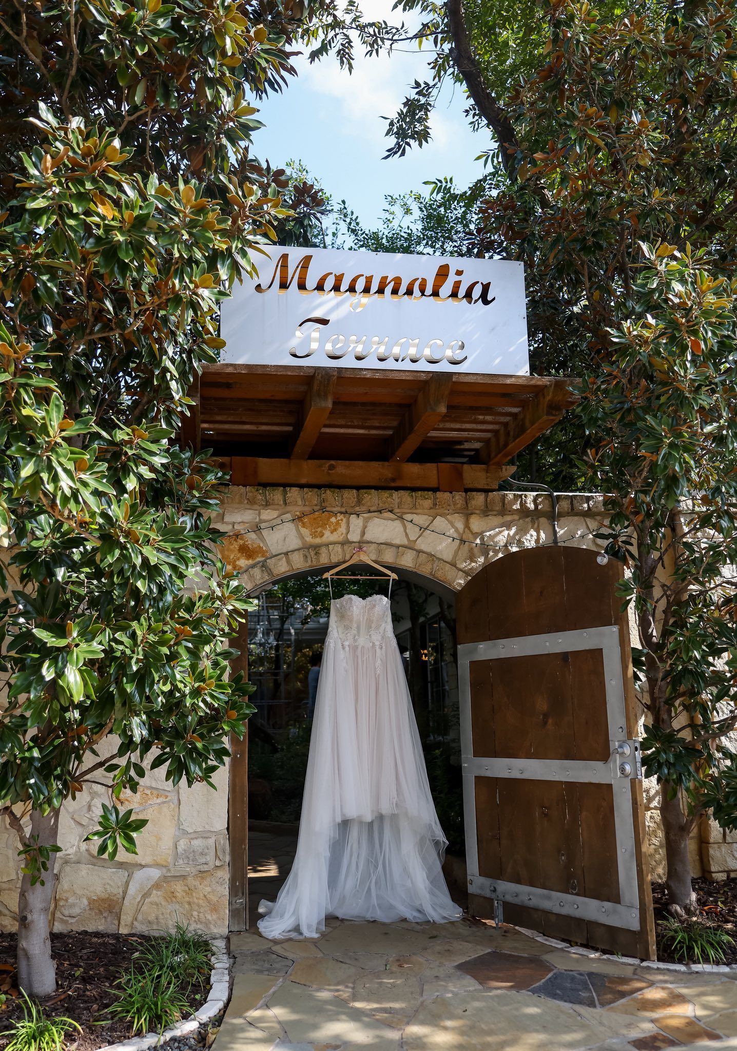 Bridal gown hanging beneath Magnolia Terrace sign at a stone arch entrance surrounded by trees.