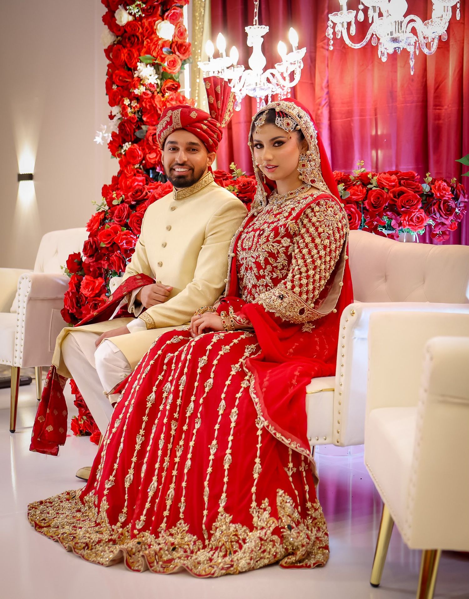 Indian bride and groom in stunning red and gold traditional wedding attire seated before floral backdrop.