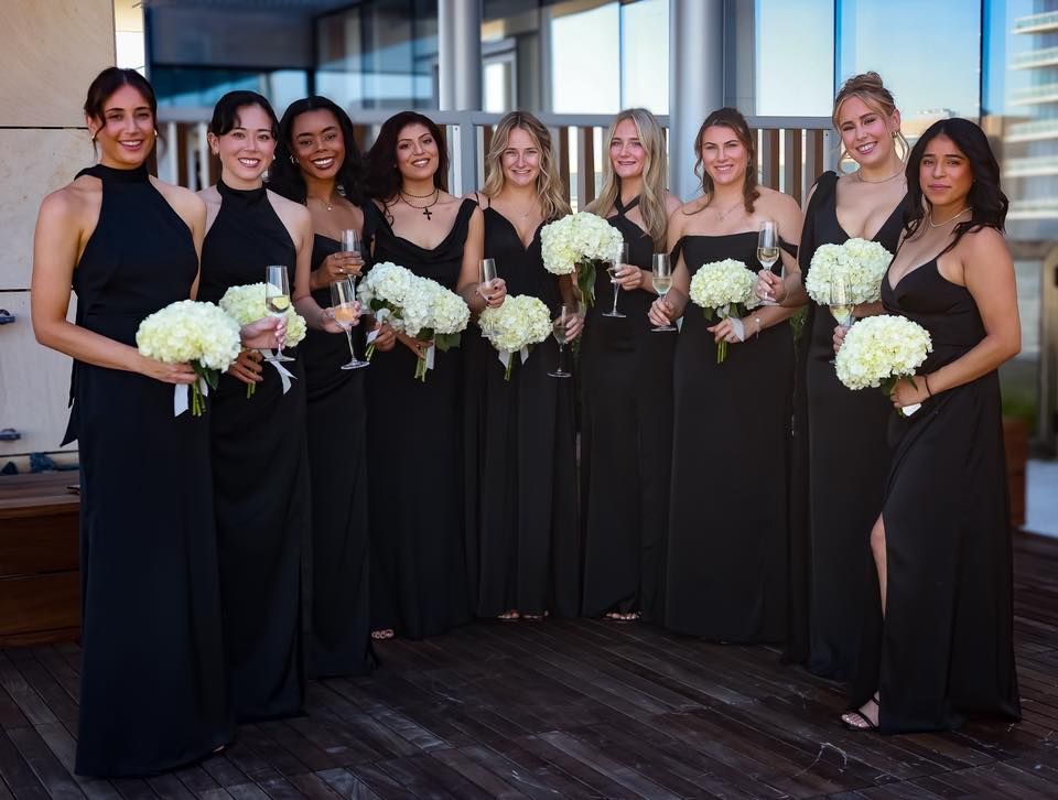 Eight bridesmaids in matching black gowns holding white hydrangea bouquets and champagne glasses at a rooftop venue.
