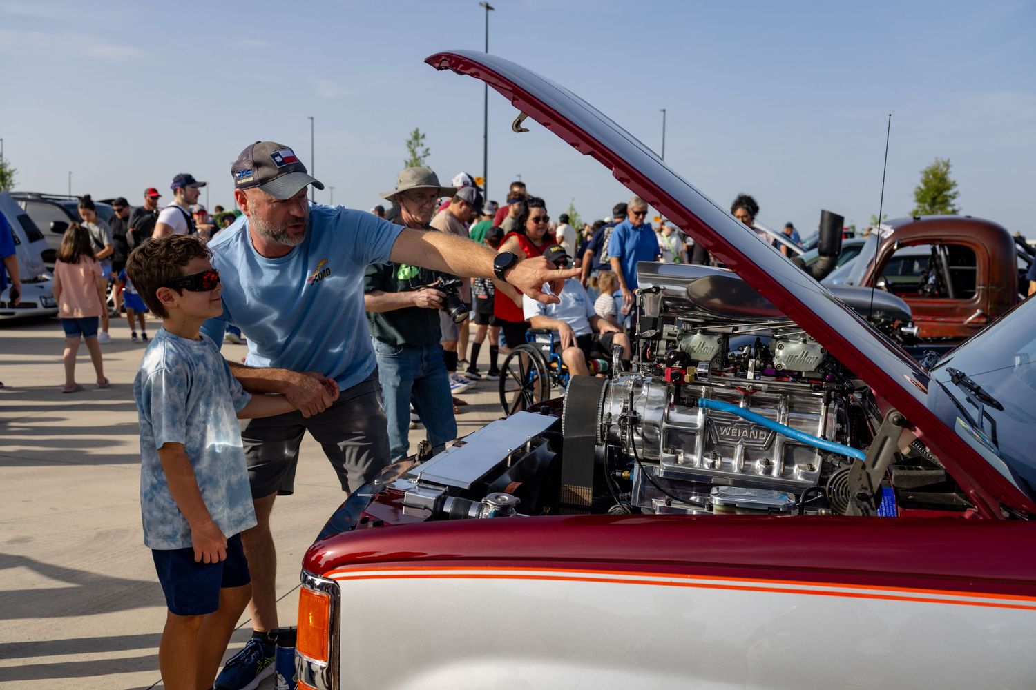 Man and child examining a classic car engine at an outdoor car show with crowd in background.