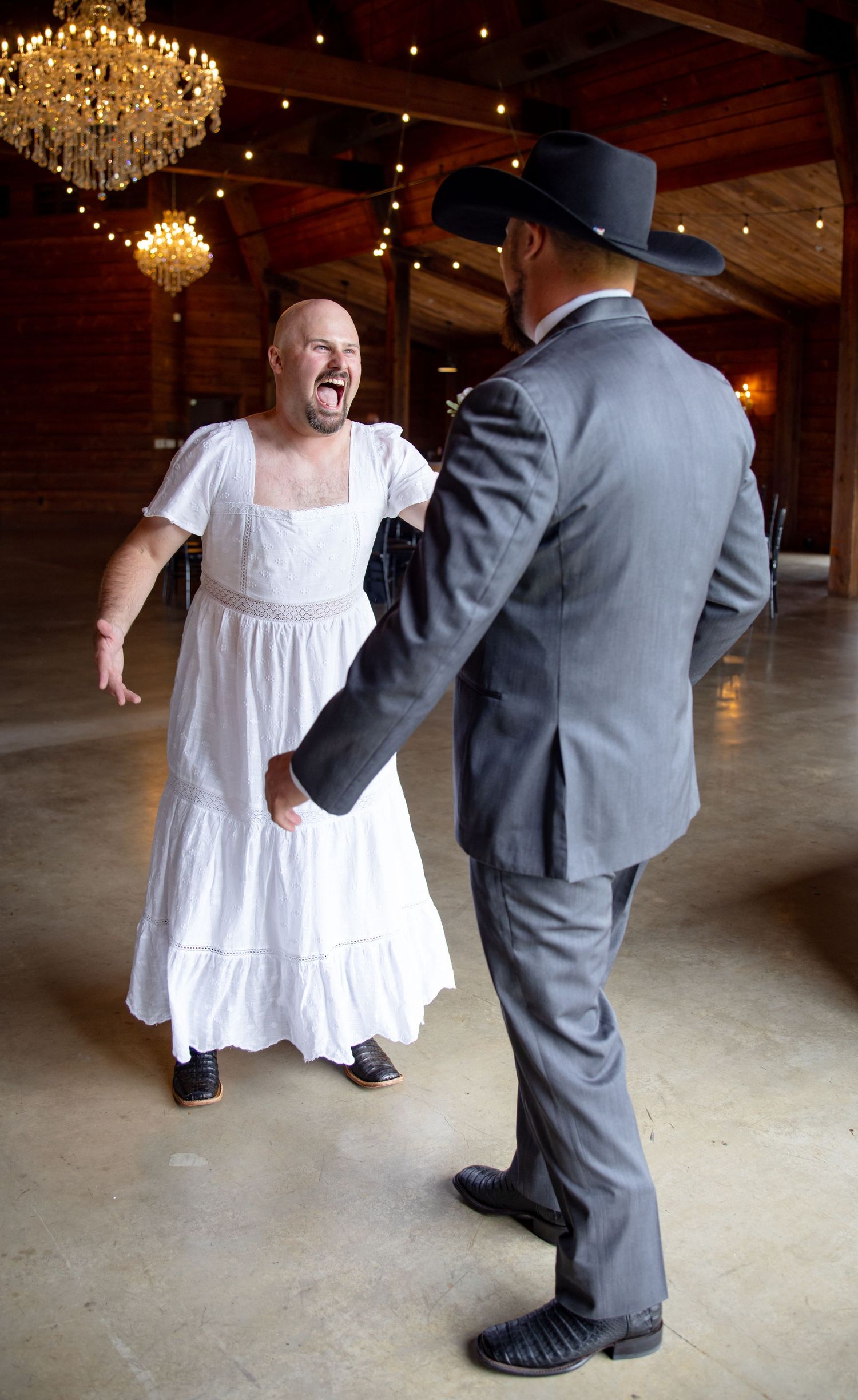 Laughing man in white dress surprises groom in gray suit and black cowboy hat inside rustic barn venue.