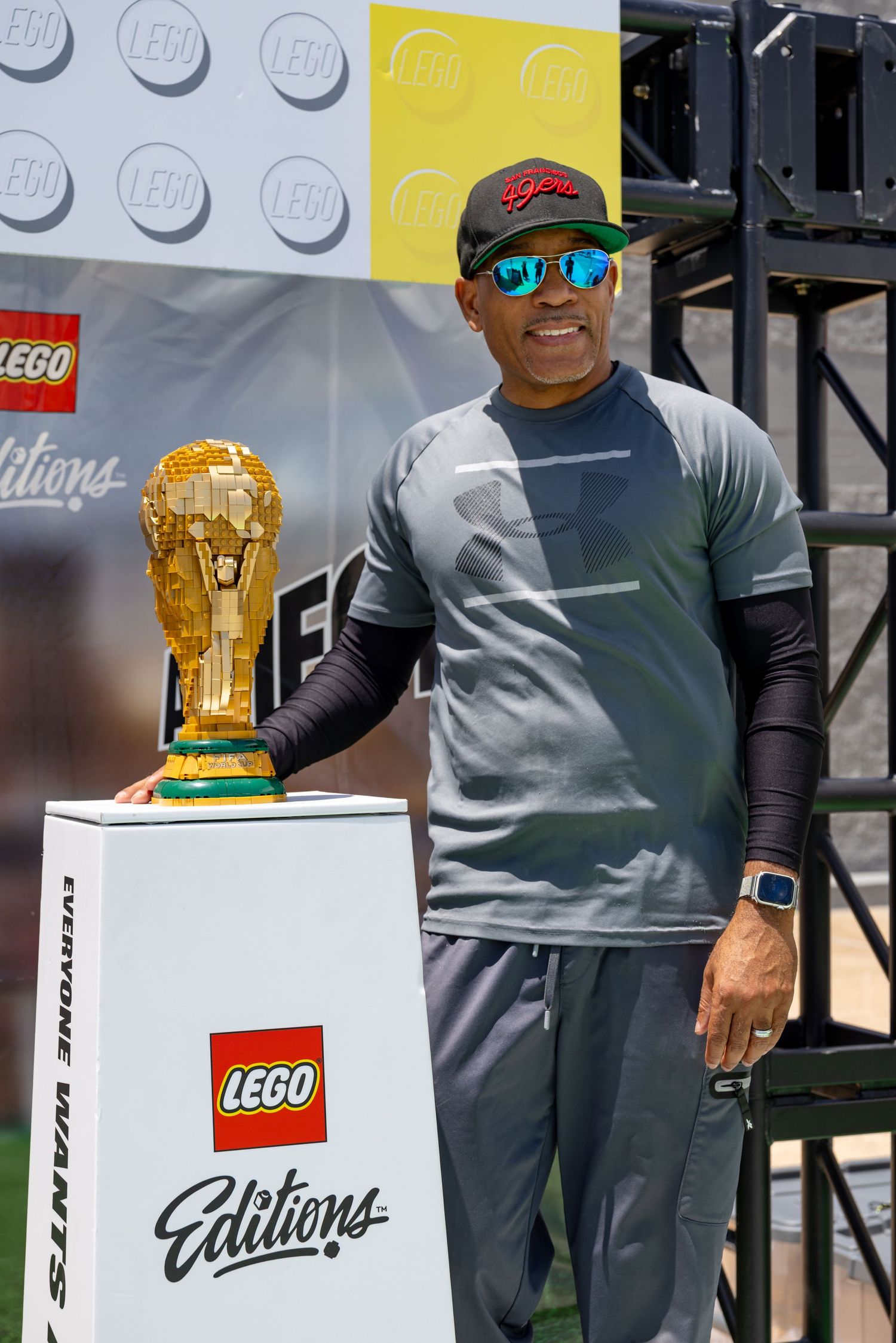 A man in a LEGO cap and sunglasses poses next to a golden LEGO World Cup trophy at an outdoor event.